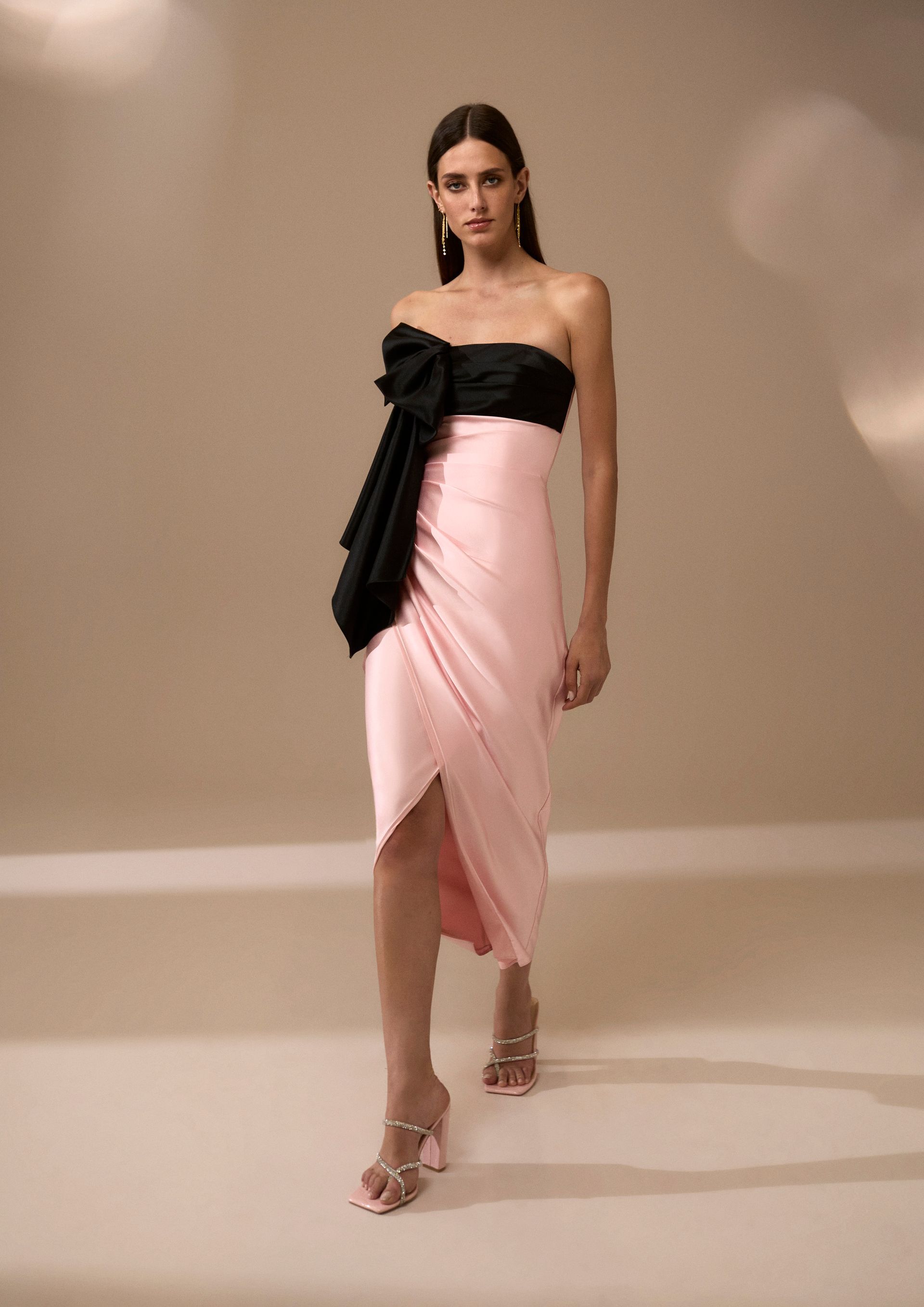 Donna in abito rosa e nero senza spalline con grande fiocco, tacchi alti, in posa in studio.