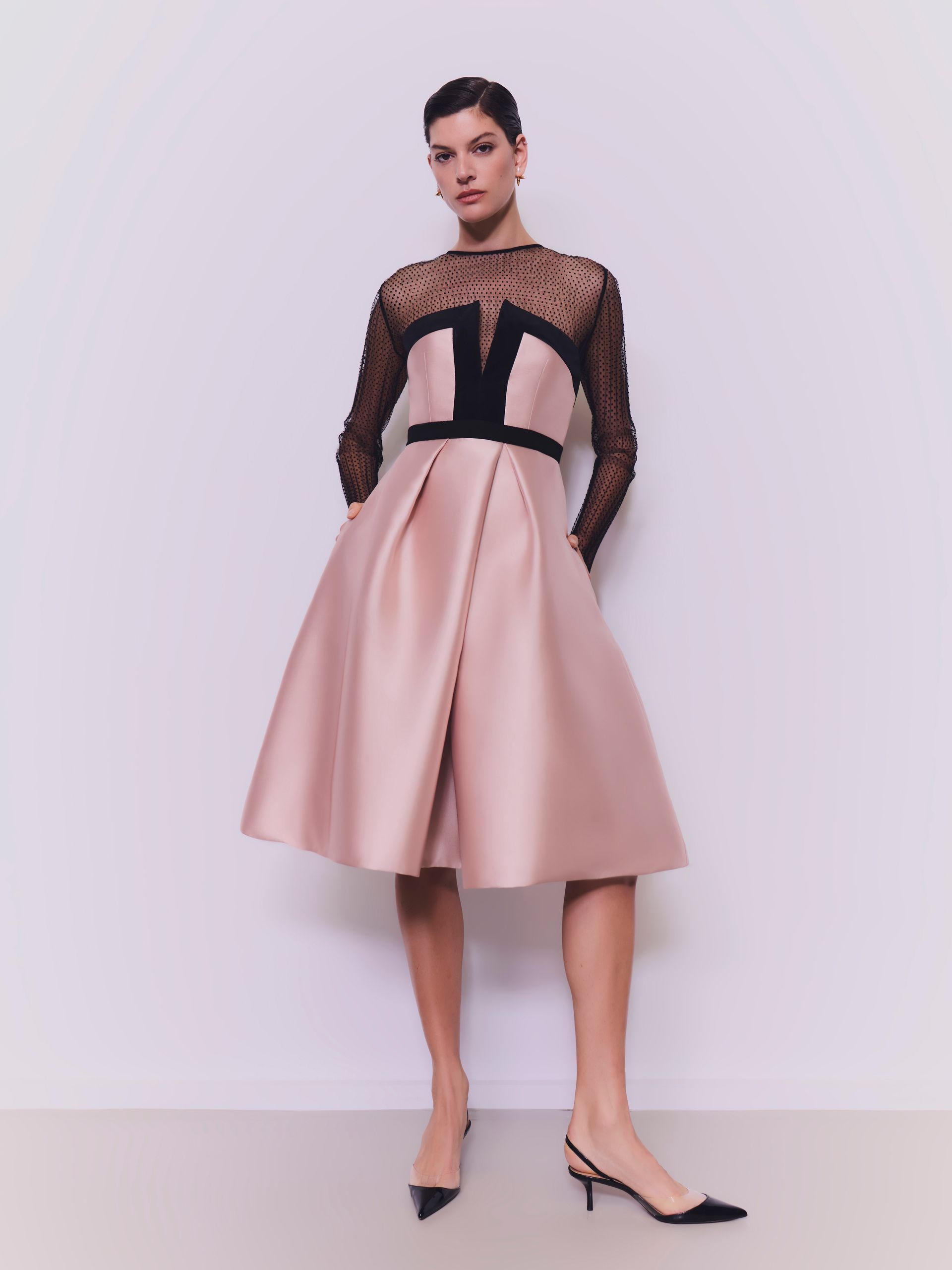 Donna che indossa un abito di raso rosa cipria con top nero trasparente e tacchi, in piedi in uno studio.