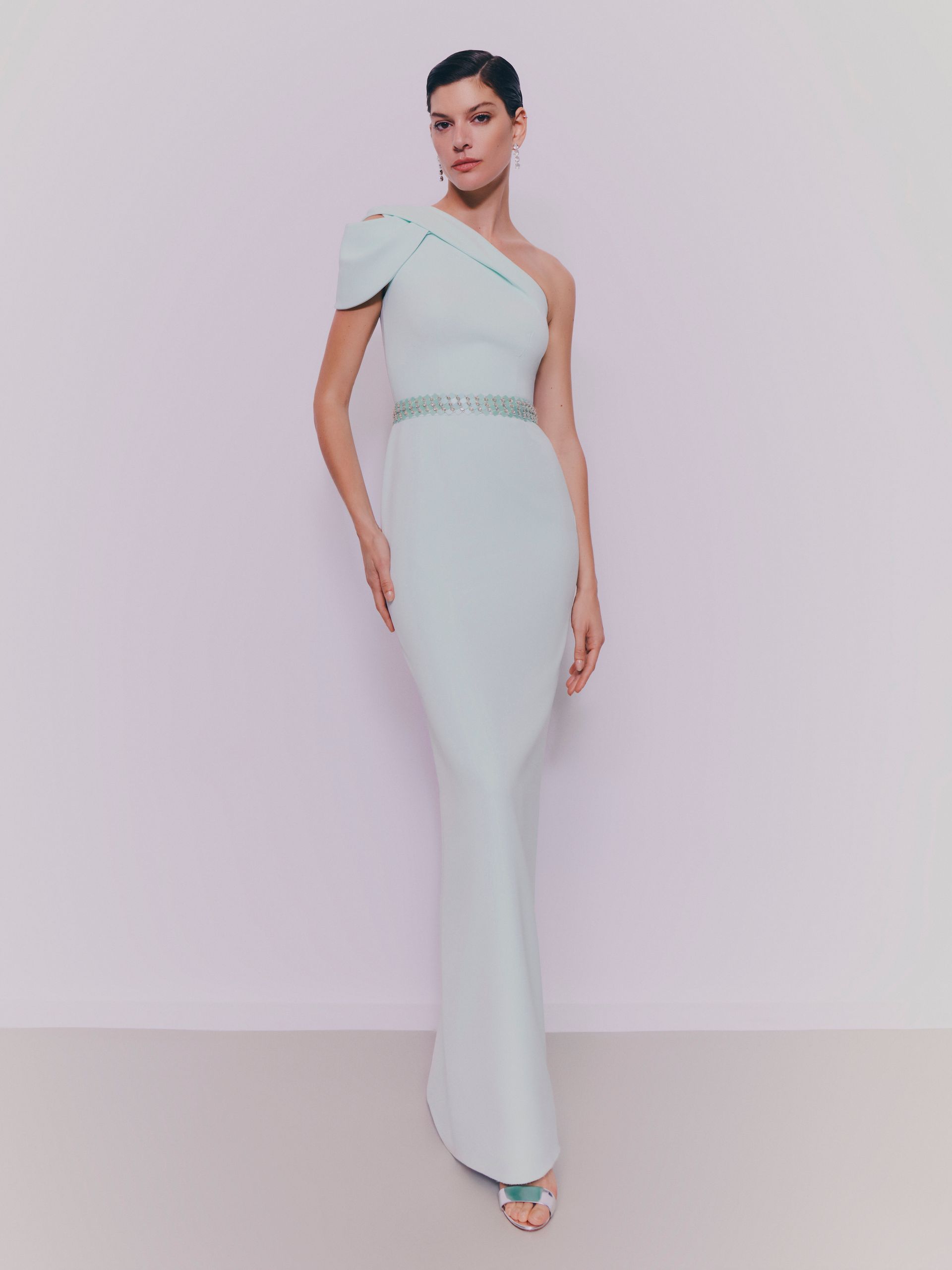Donna in abito azzurro chiaro monospalla con cintura di perline, in piedi, con lo sguardo rivolto in avanti.