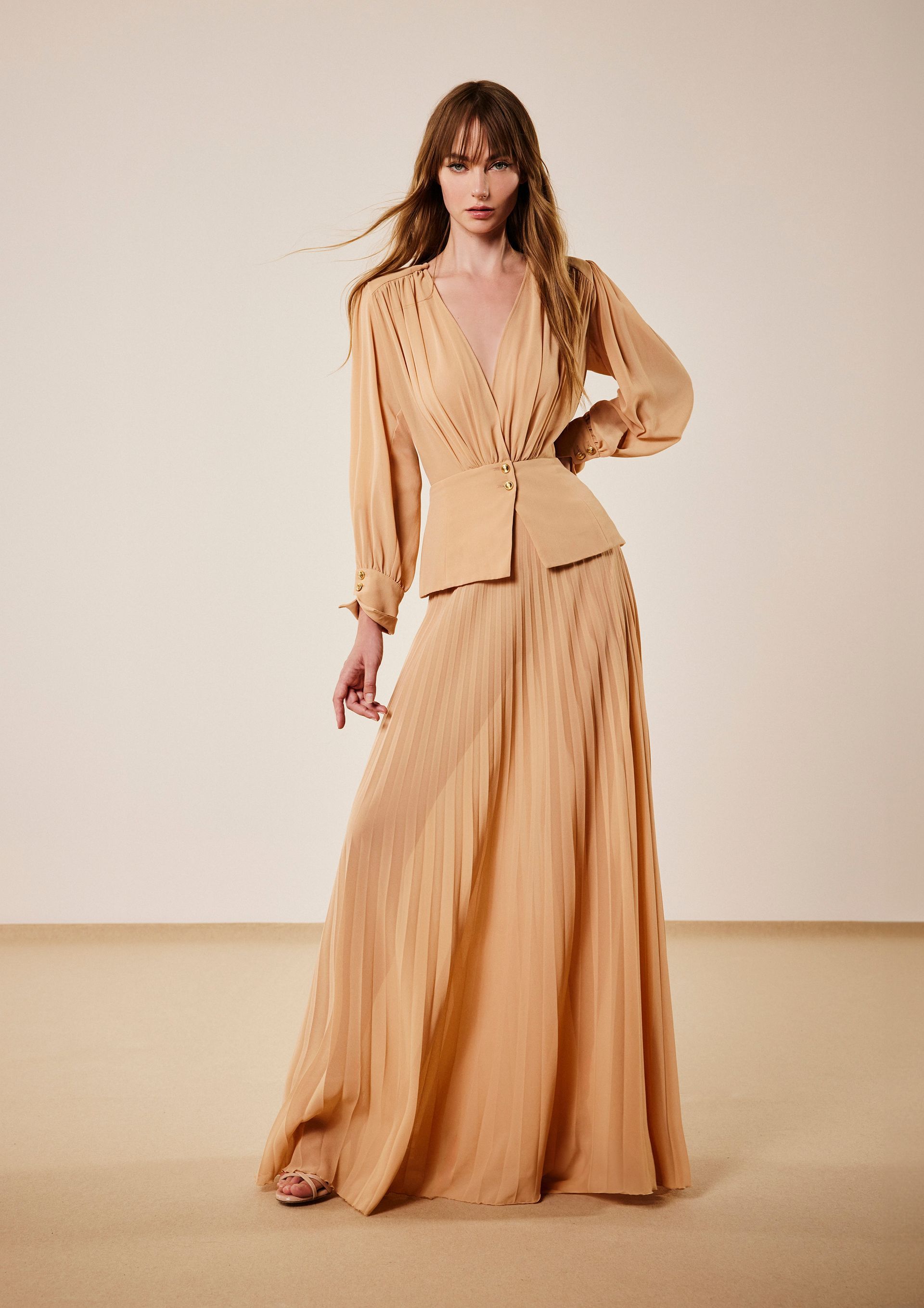 Donna in un abito svolazzante color pesca con maniche lunghe e gonna plissettata, in posa in studio.