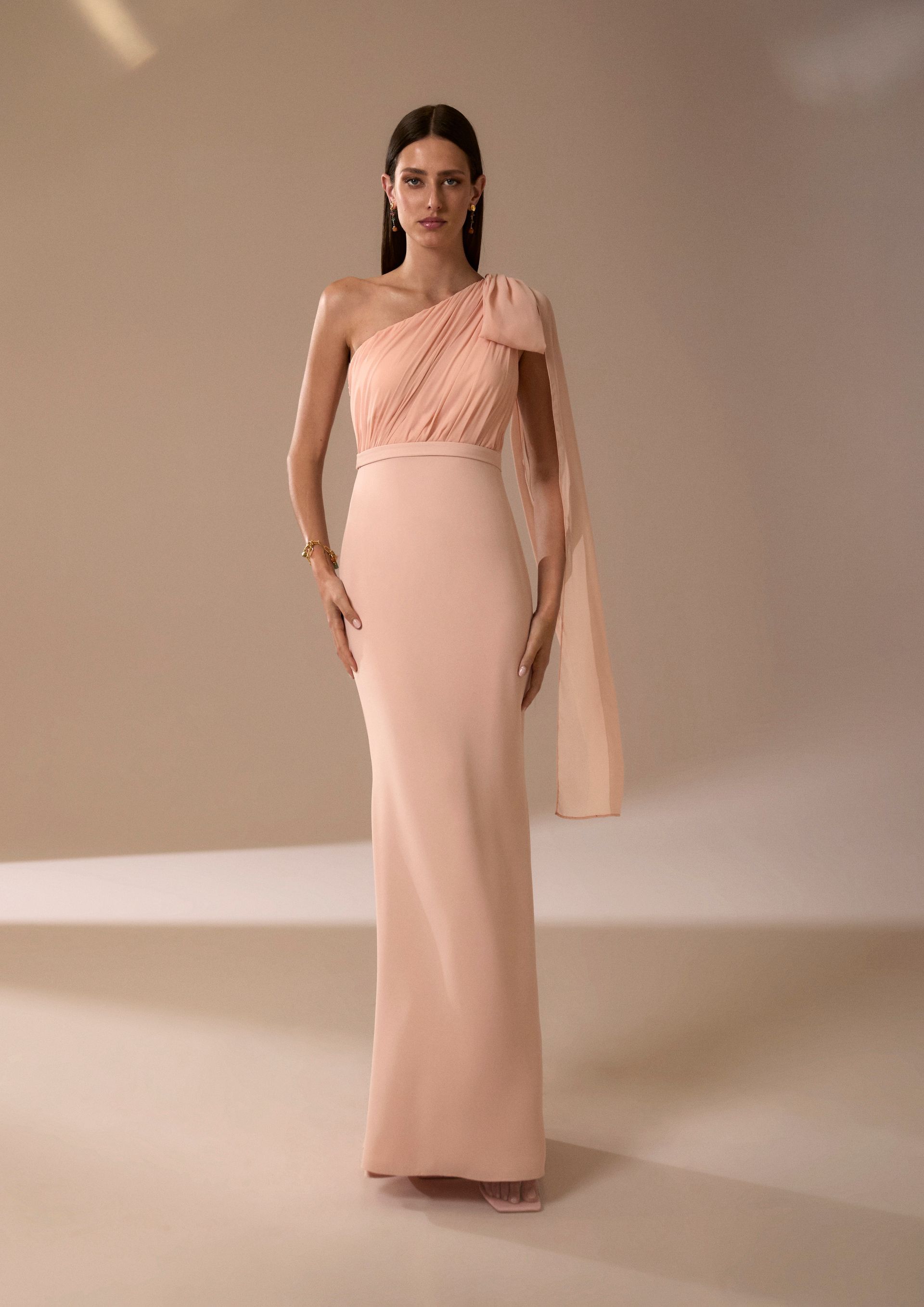 Donna in abito monospalla color pesca con sciarpa attaccata, in piedi in studio.