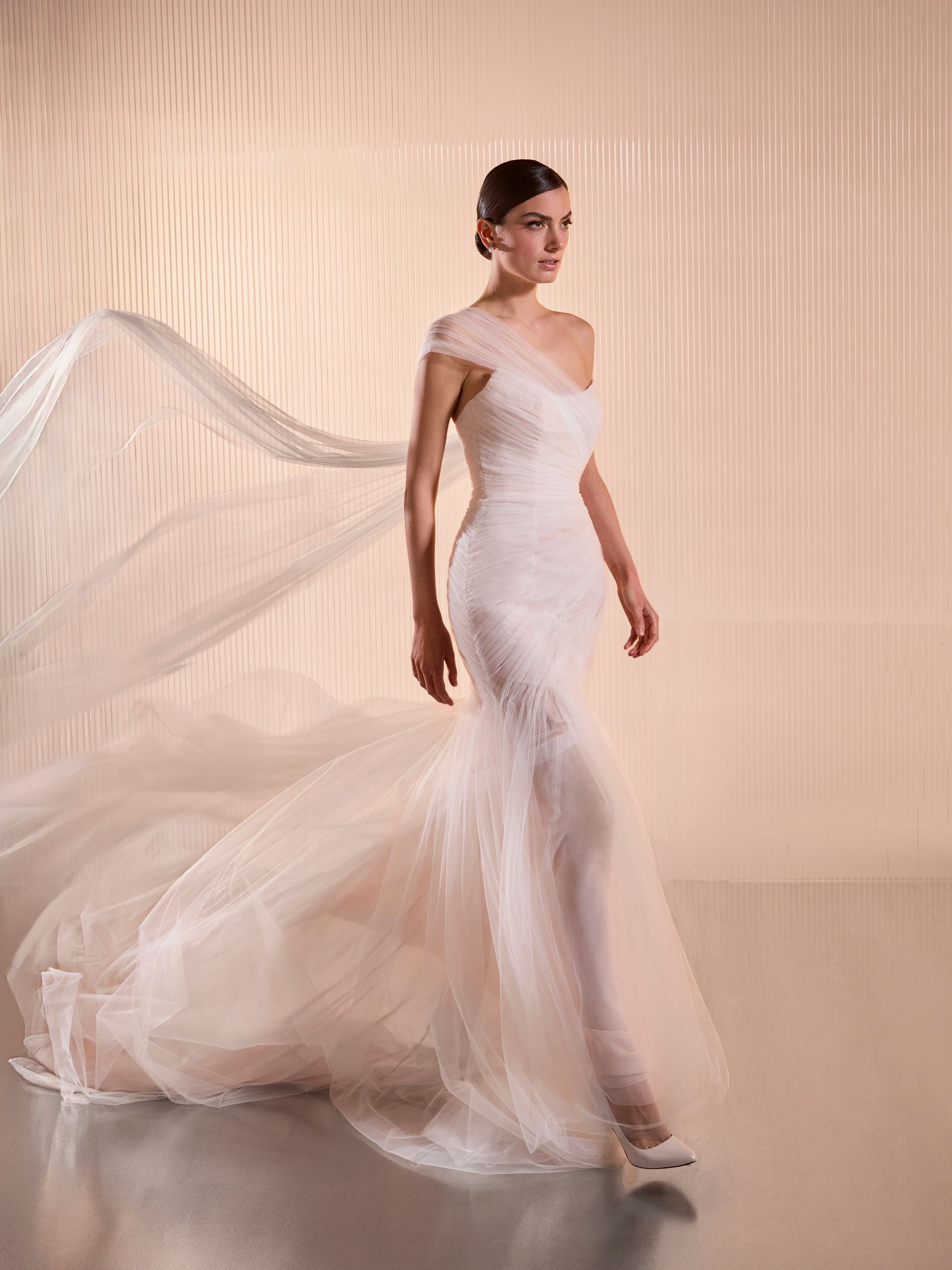 Donna in abito da sposa bianco monospalla, strascico fluente. Cammina, espressione neutra, sfondo color pesca.