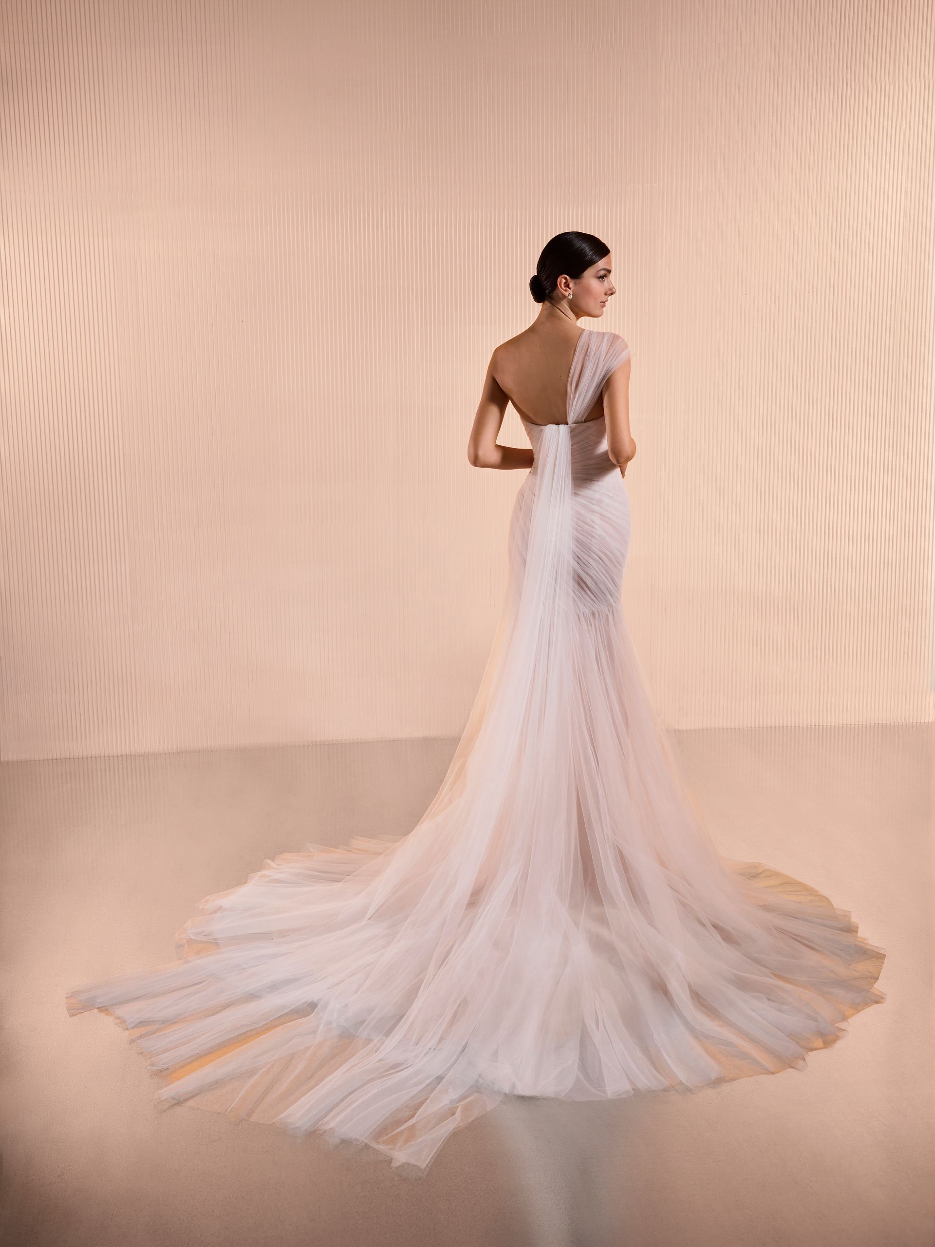 Donna in abito da sposa bianco con lungo strascico, in piedi su uno sfondo color pesca.