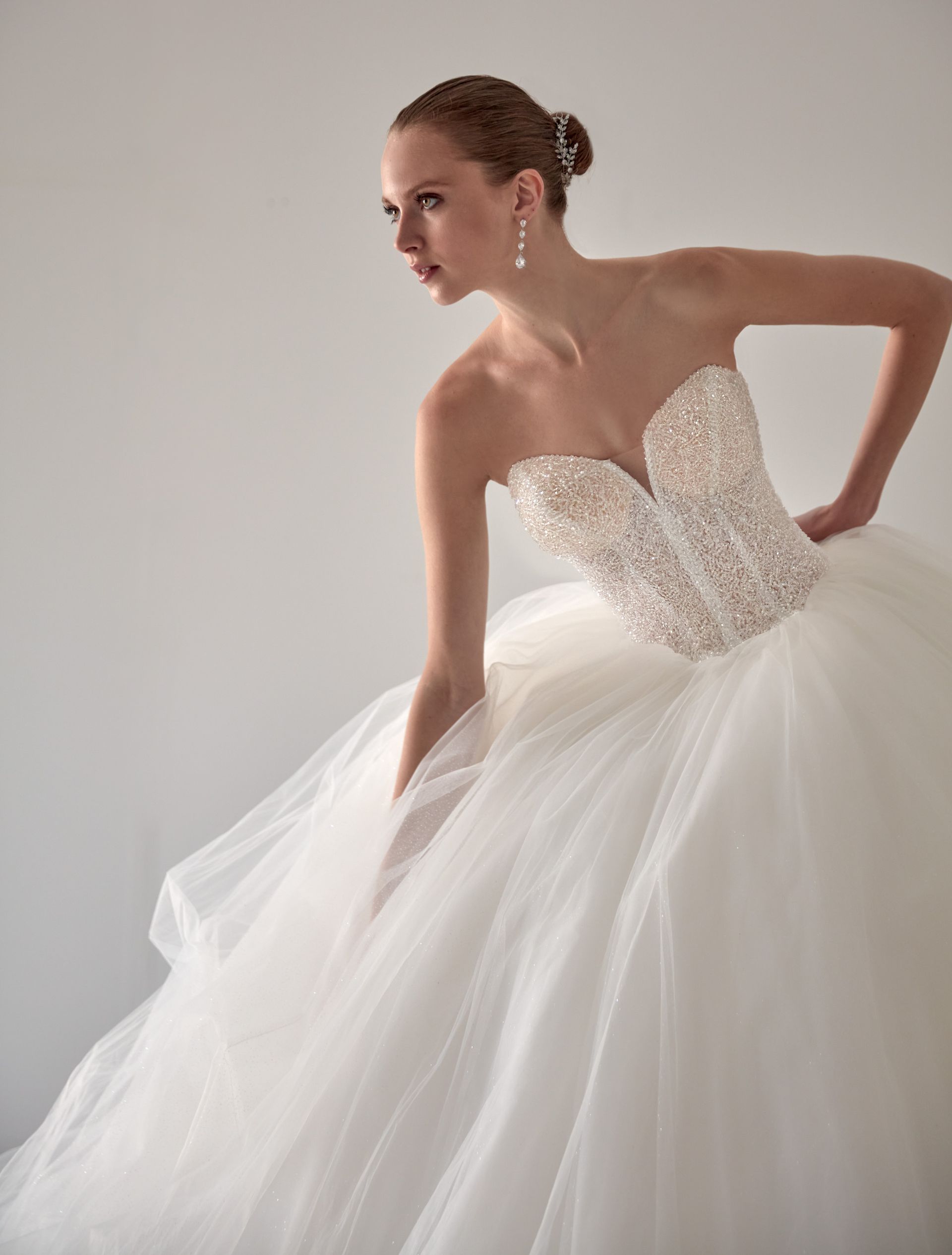 Donna in abito da sposa bianco senza spalline, corpetto decorato, gonna in tulle, in posa su uno sfondo bianco.