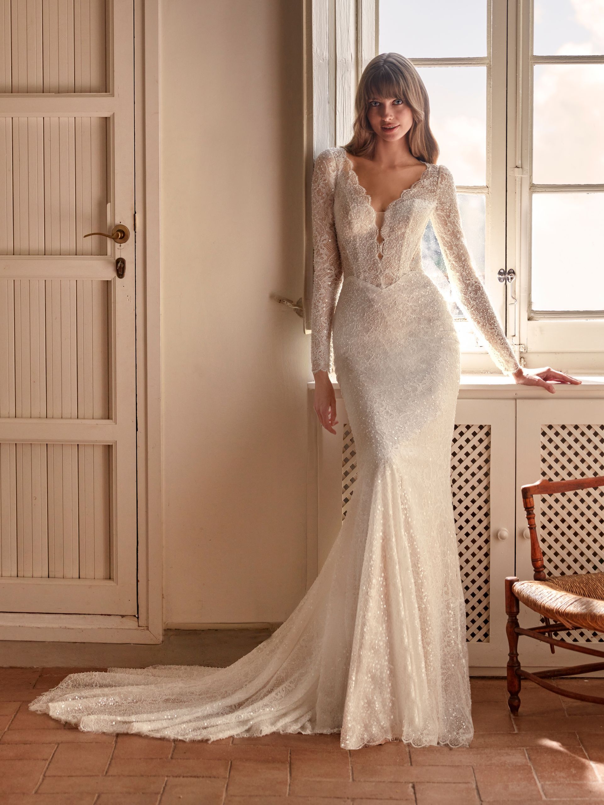 Una donna con un abito da sposa con perline e maniche lunghe è in piedi vicino a una finestra in una stanza