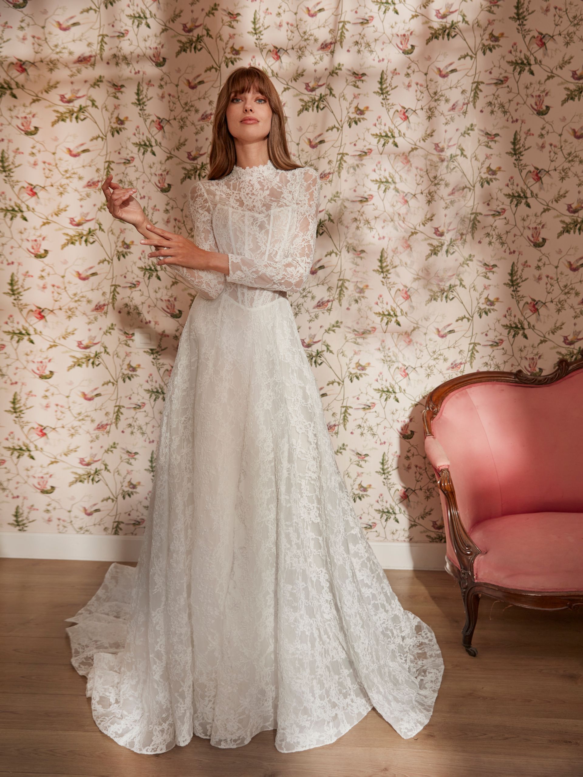 Una donna in abito di pizzo bianco è in piedi contro una carta da parati floreale, accanto a un divano rosa.