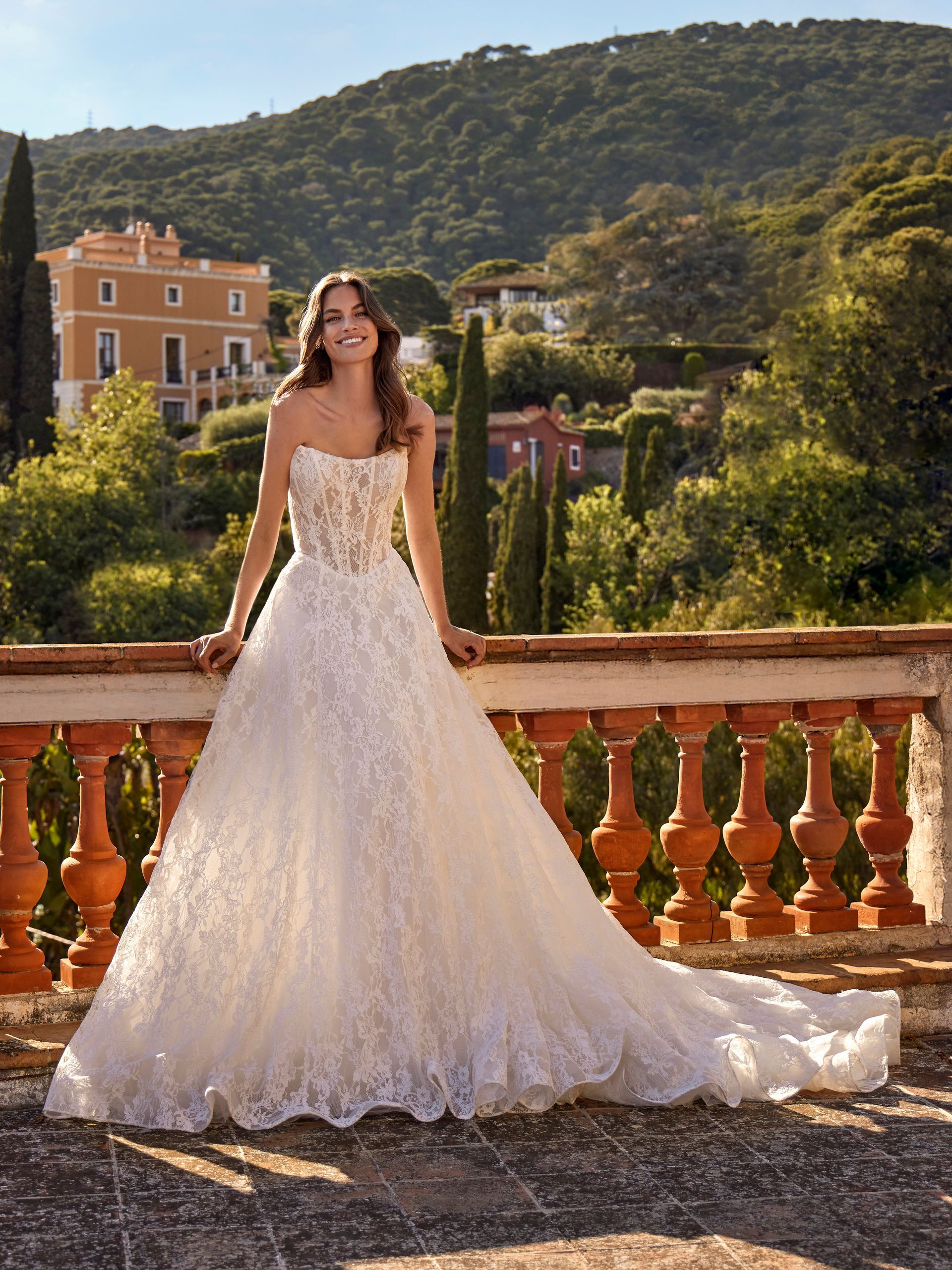 Donna in abito da sposa bianco senza spalline, sorridente all'aperto. È appoggiata a un balcone in pietra 