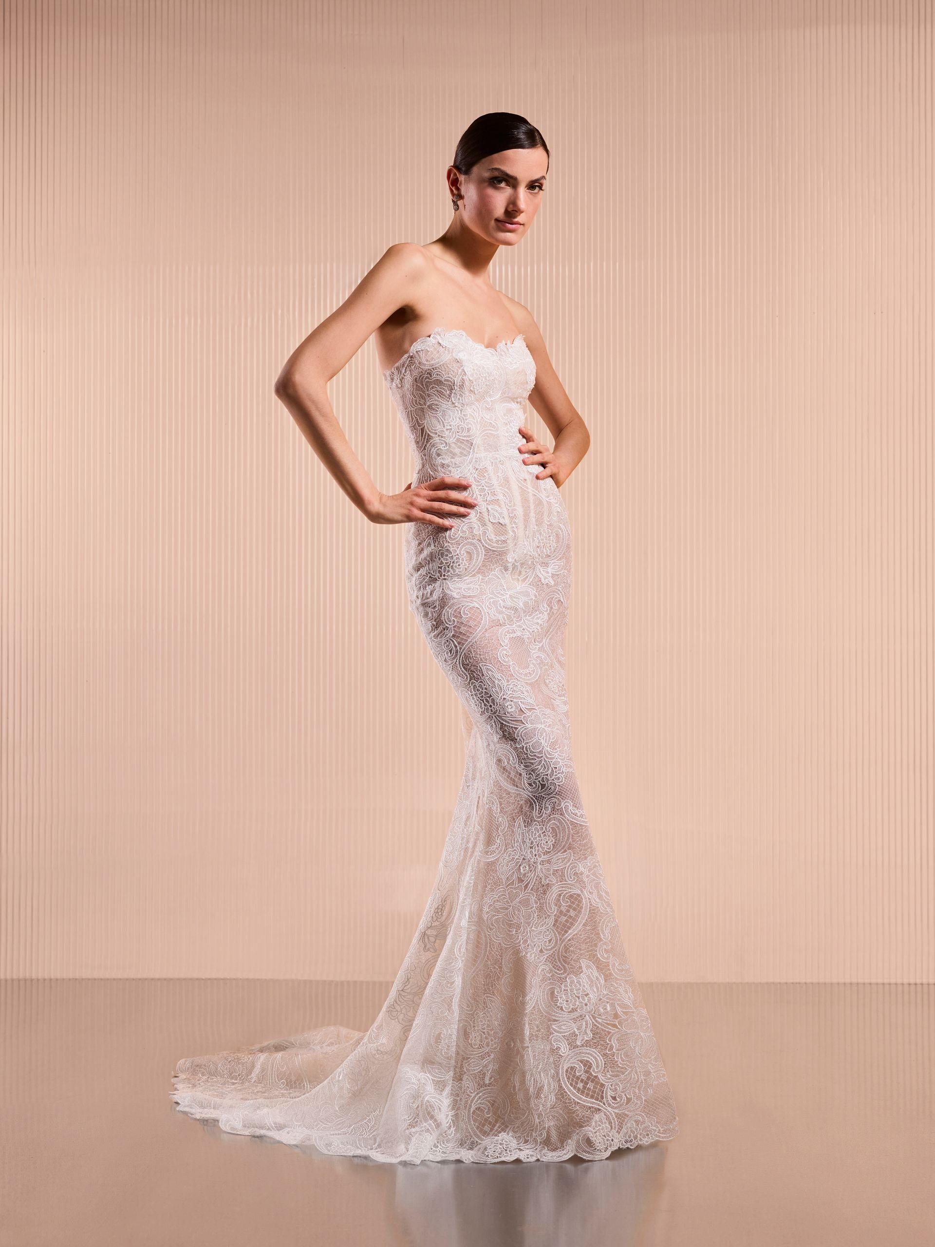 Donna in abito bianco a sirena decorato e senza spalline, mani sui fianchi, in posa su uno sfondo chiaro.