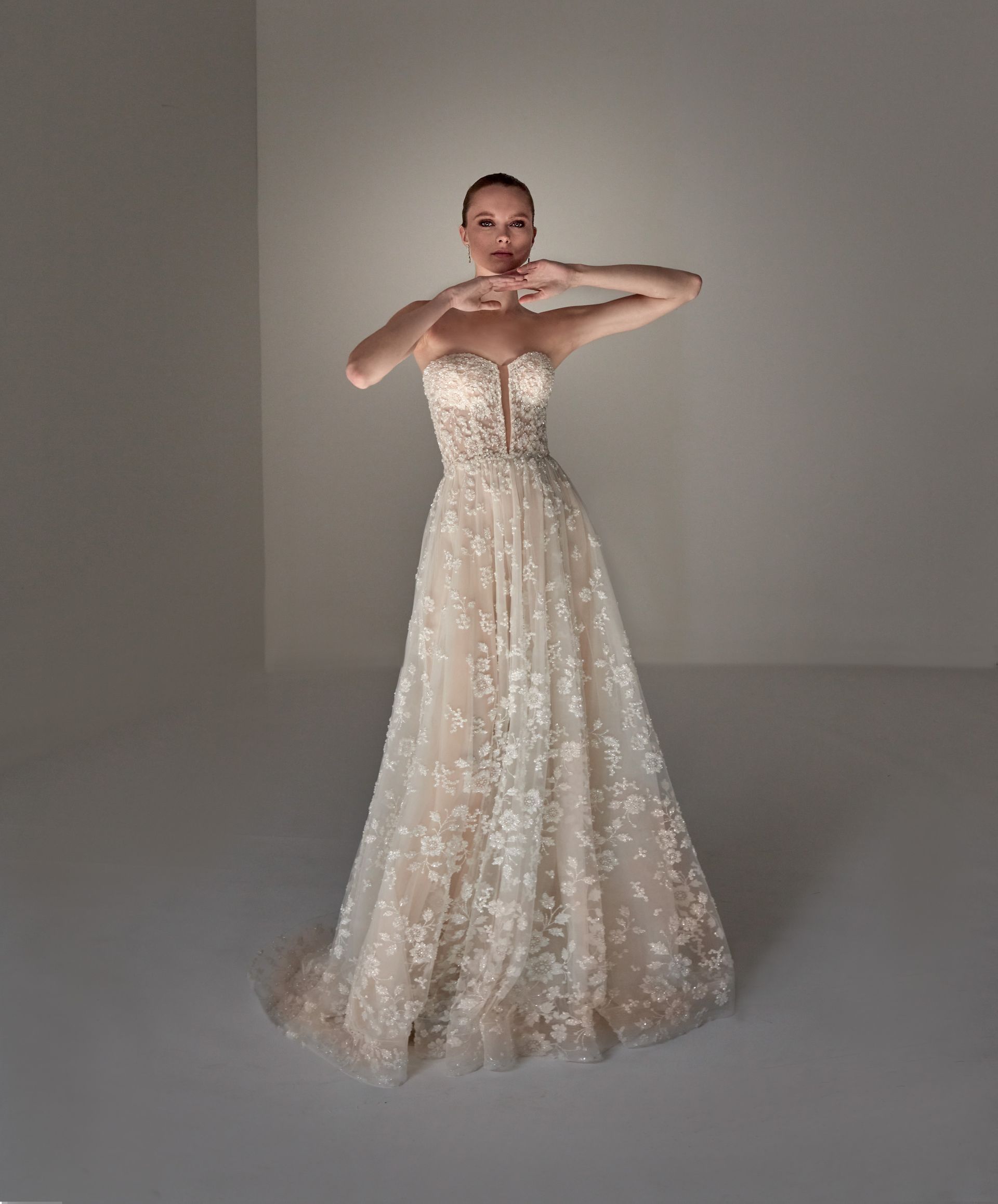 Donna in abito da sposa senza spalline, braccia alzate, mani vicino al viso. Abito beige chiaro con dettagli floreali.