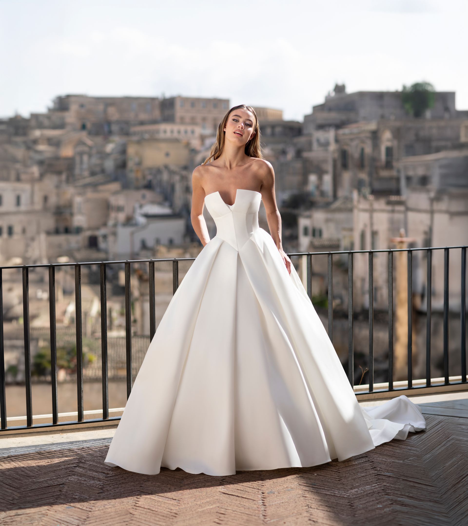 Donna in abito da sposa bianco senza spalline, in piedi all'aperto con una città antica sullo sfondo.