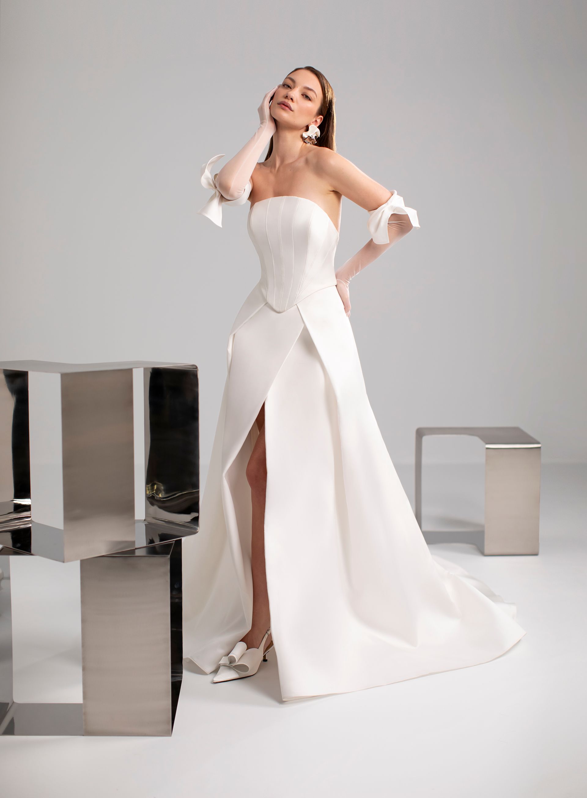 Donna in abito da sposa bianco senza spalline con spacco profondo, in posa accanto a moderni blocchi di metallo.