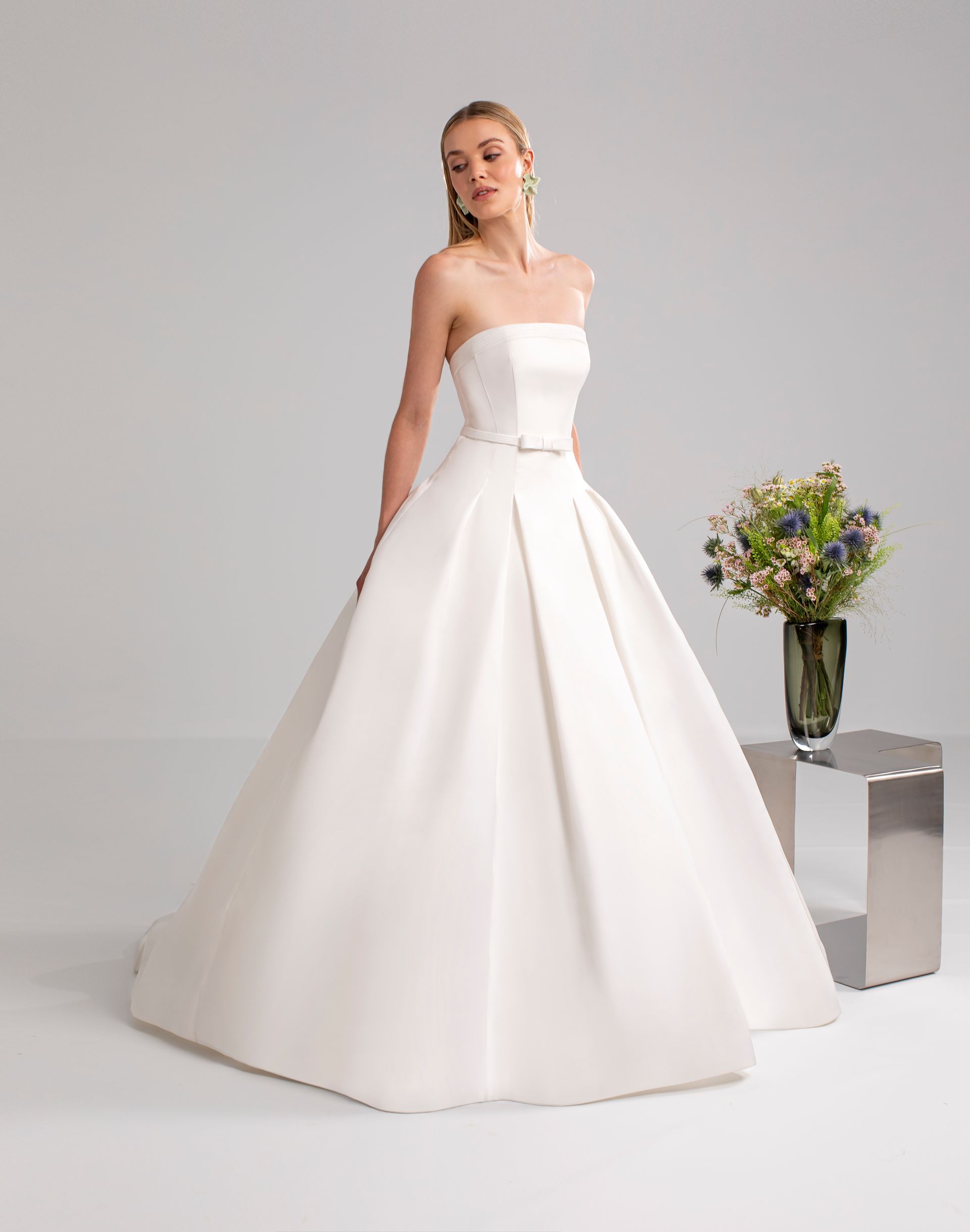 Donna in abito da sposa bianco senza spalline, in piedi accanto a un vaso di fiori su un tavolino argentato.