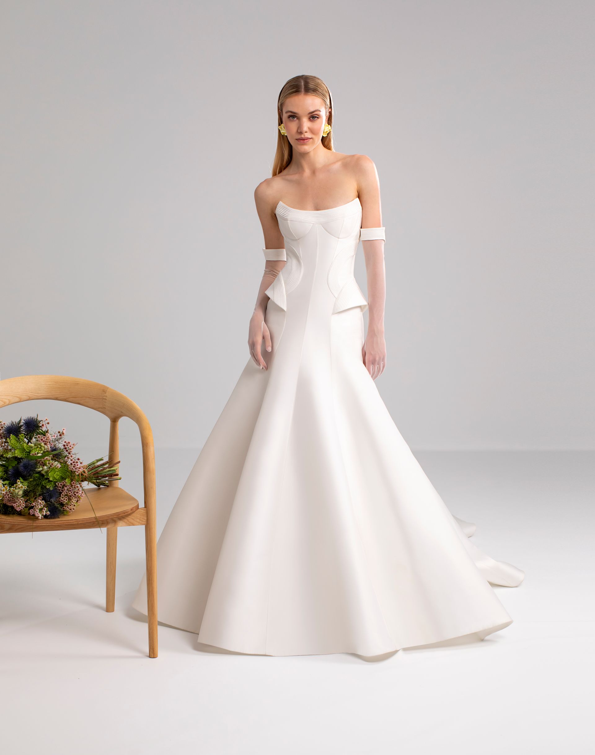 Donna sorridente in abito da sposa bianco senza spalline, accanto a una sedia di legno con dei fiori.