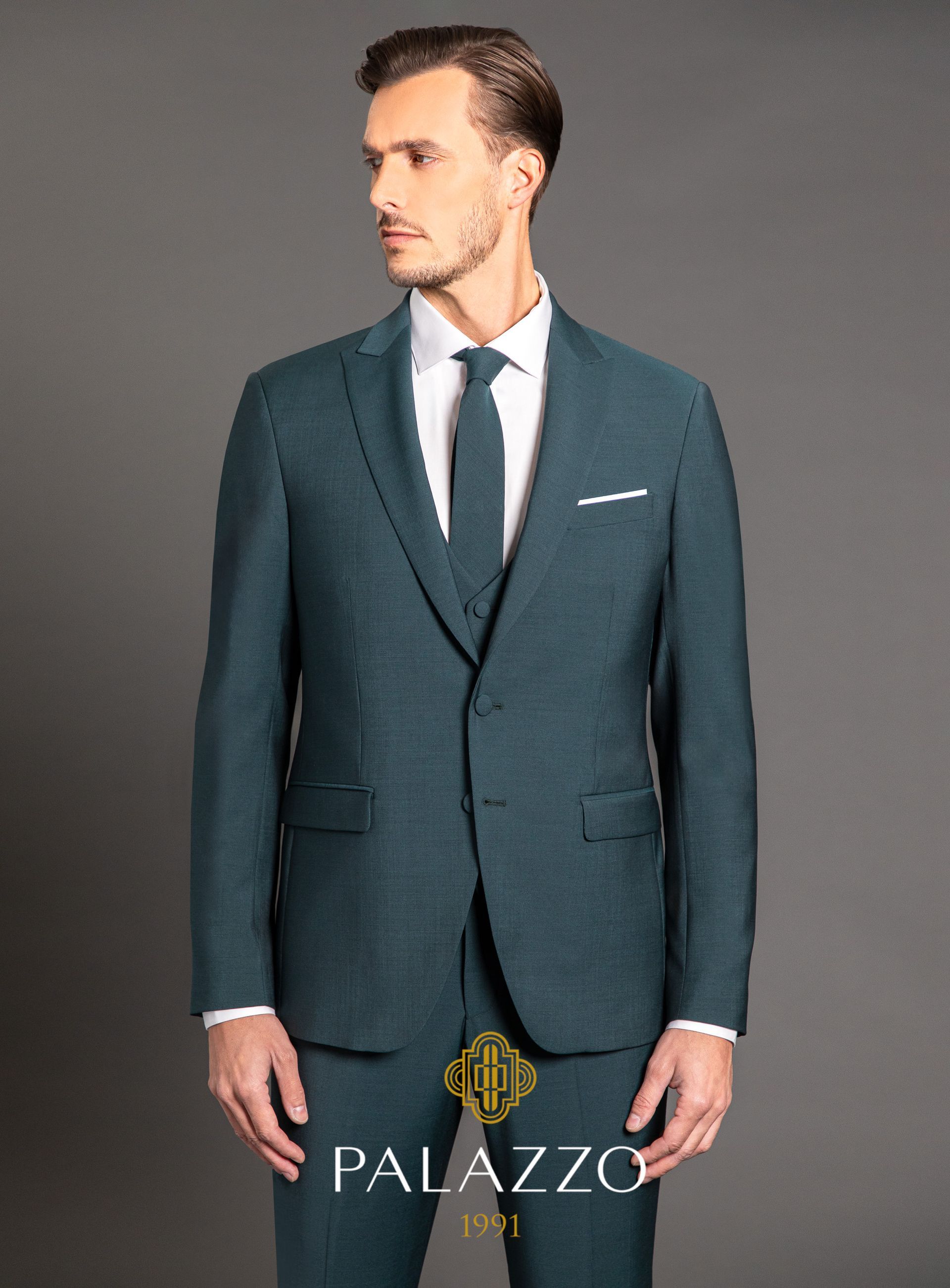 Uomo in abito verde acqua e cravatta, camicia bianca, in posa, sfondo grigio.