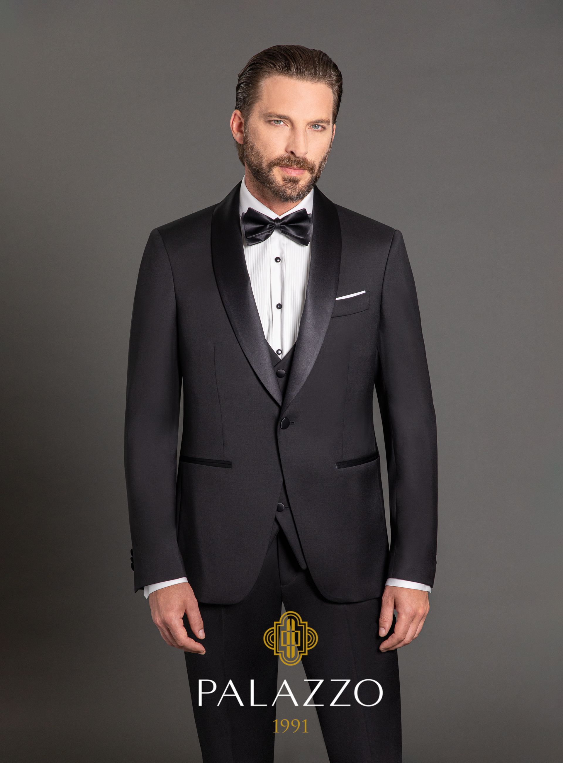 Uomo in smoking nero, papillon, camicia bianca e gilet, in piedi su uno sfondo grigio. Logo Palazzo.