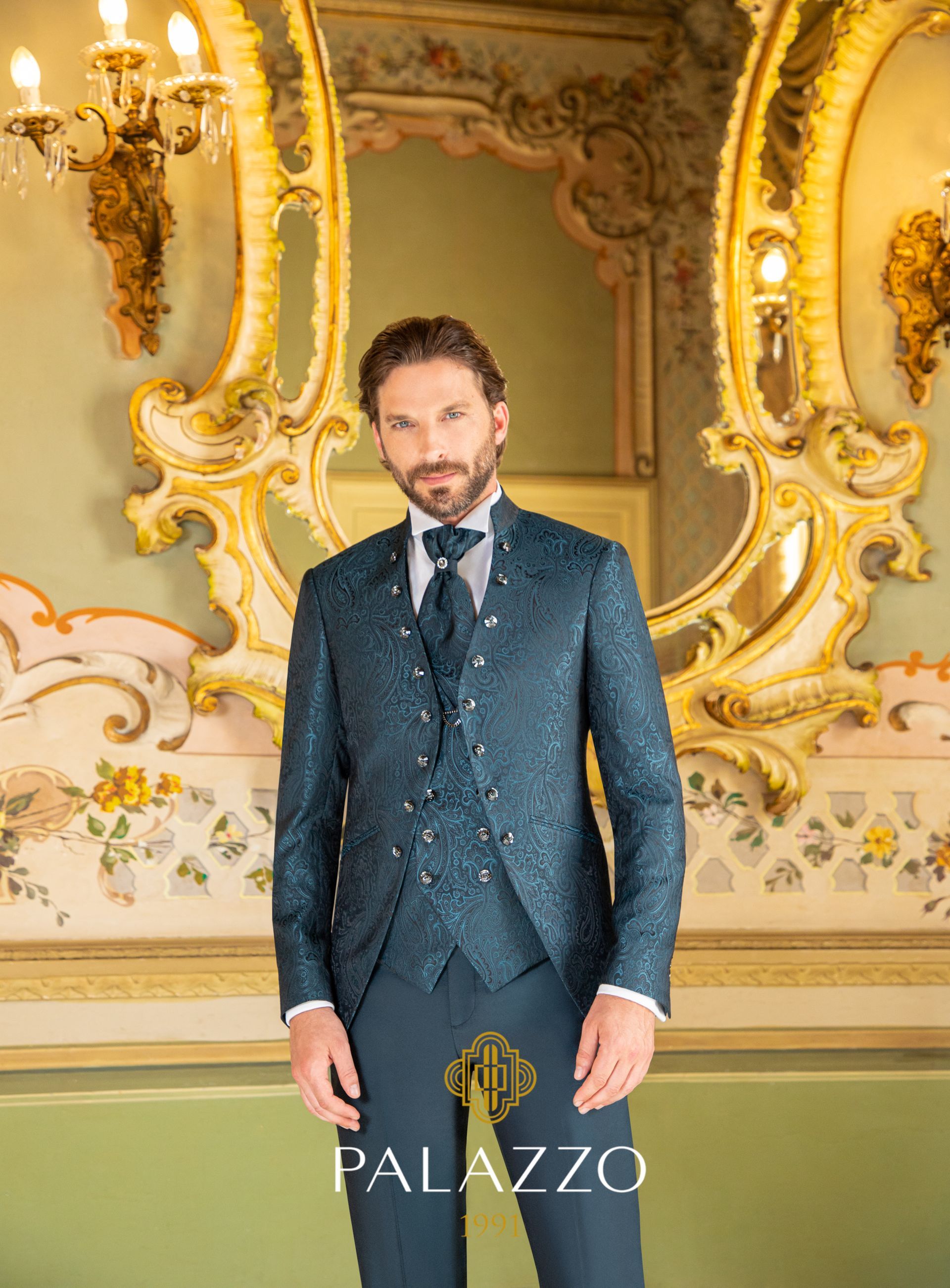 Un uomo in un elaborato abito color verde acqua è in piedi all'interno, accanto a uno specchio dorato decorativo.