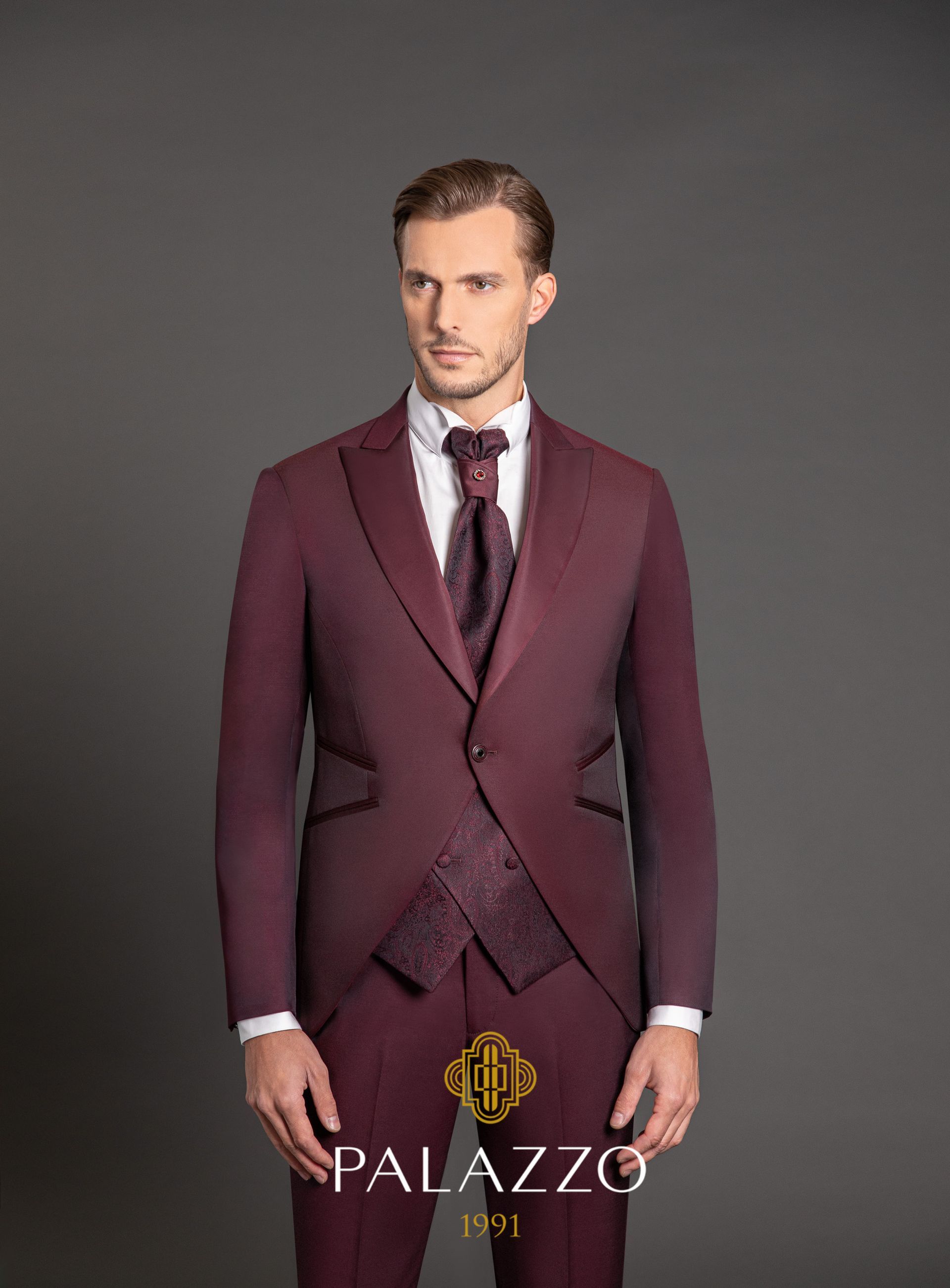 Uomo in abito bordeaux, camicia bianca e cravatta, in piedi davanti a uno sfondo grigio.