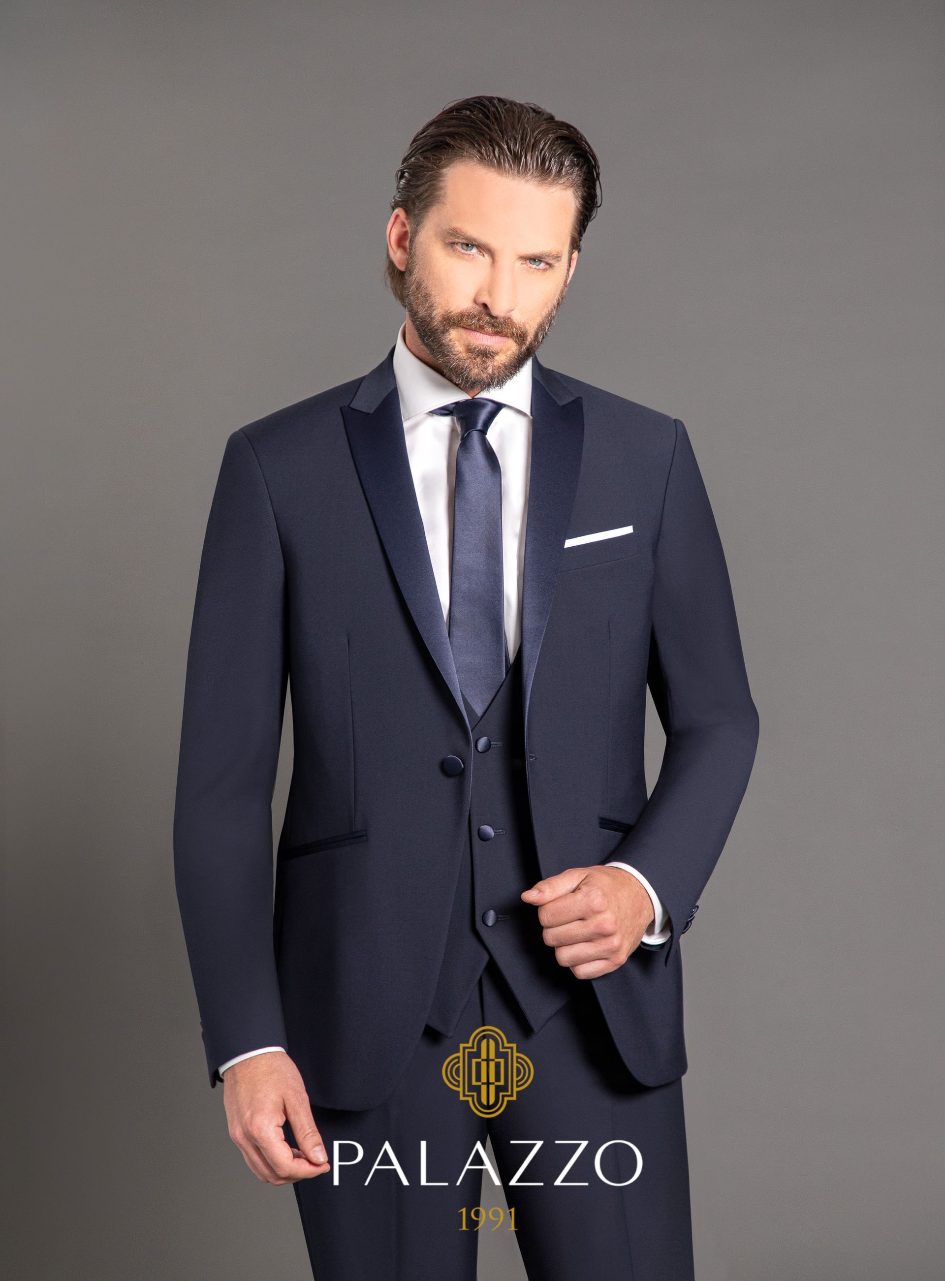 Un uomo in smoking blu scuro, con un leggero sorriso, in piedi davanti a uno sfondo grigio.