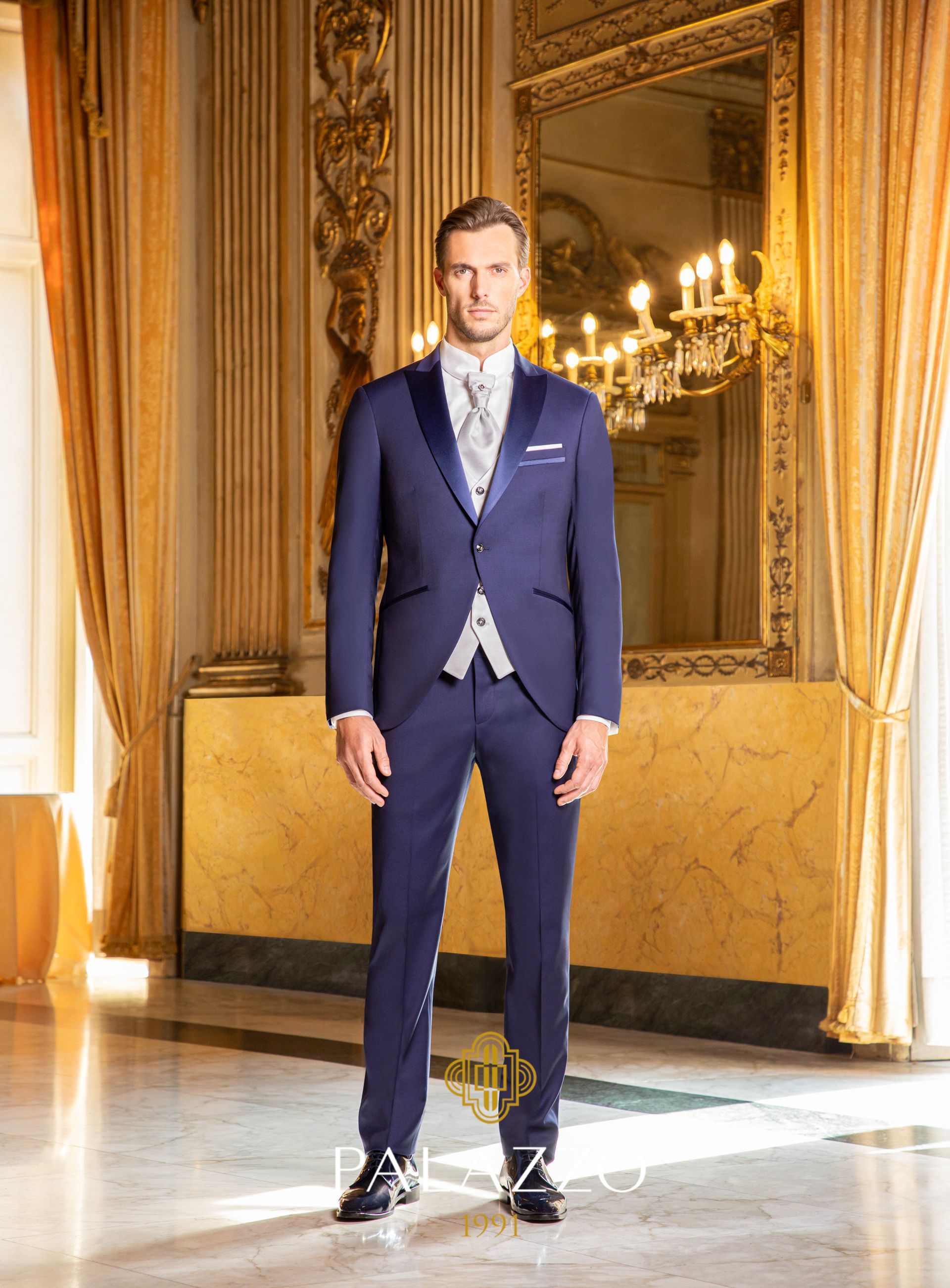 Un uomo in abito blu navy e gilet, in piedi in una stanza elegante con un grande specchio e un lampadario.