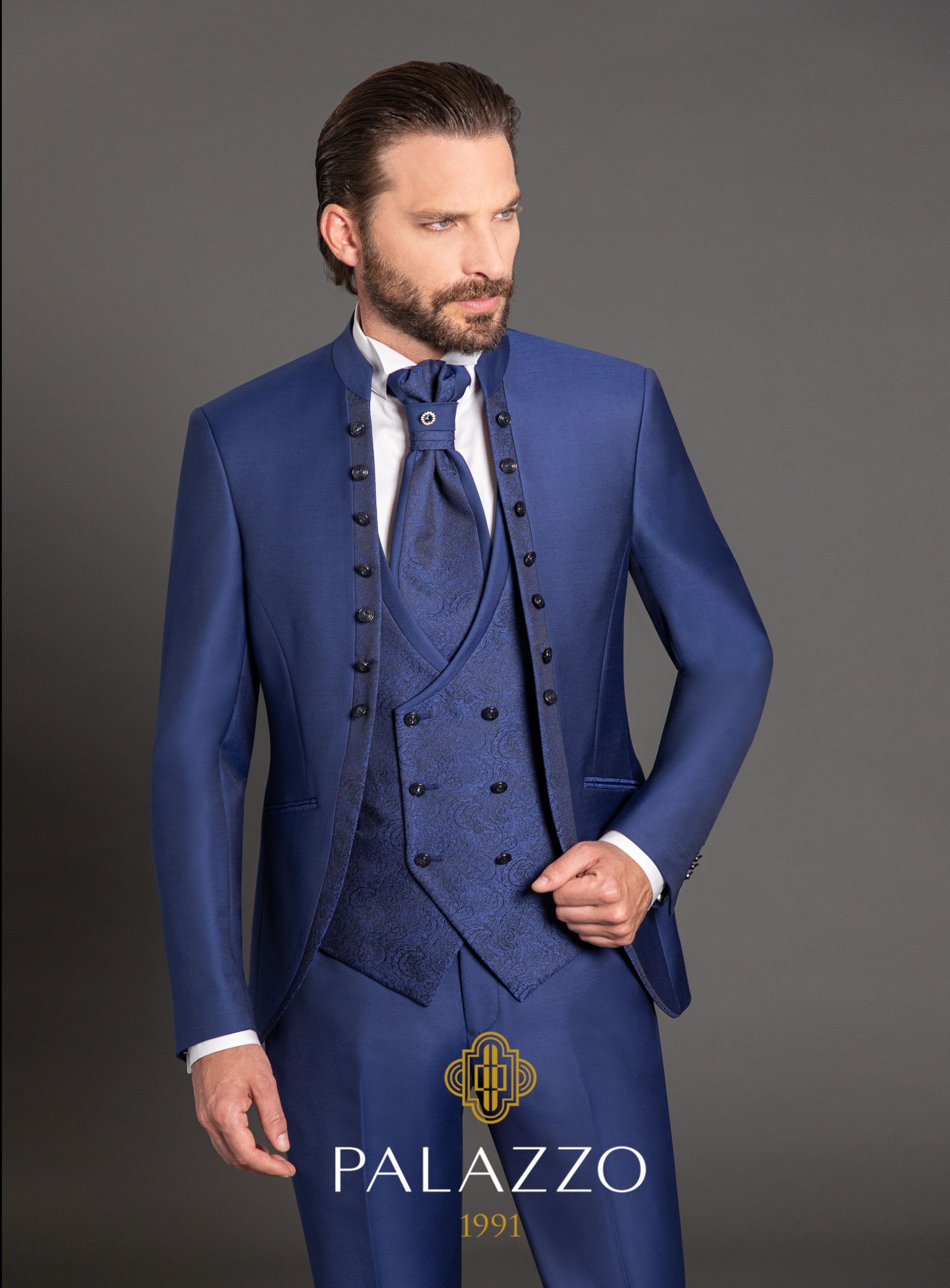 Uomo in abito formale blu con bottoni decorati, in piedi davanti a uno sfondo grigio.