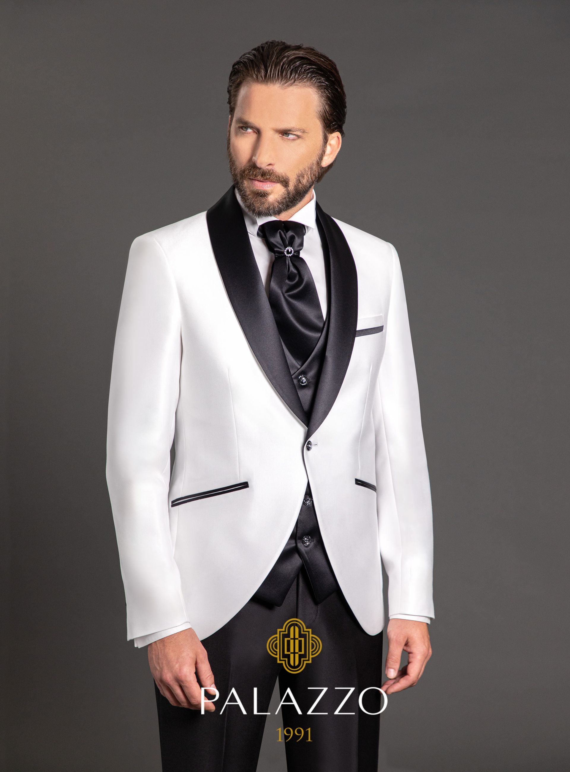 Uomo in smoking bianco con risvolti neri, cravatta e gilet neri, in posa su uno sfondo grigio.
