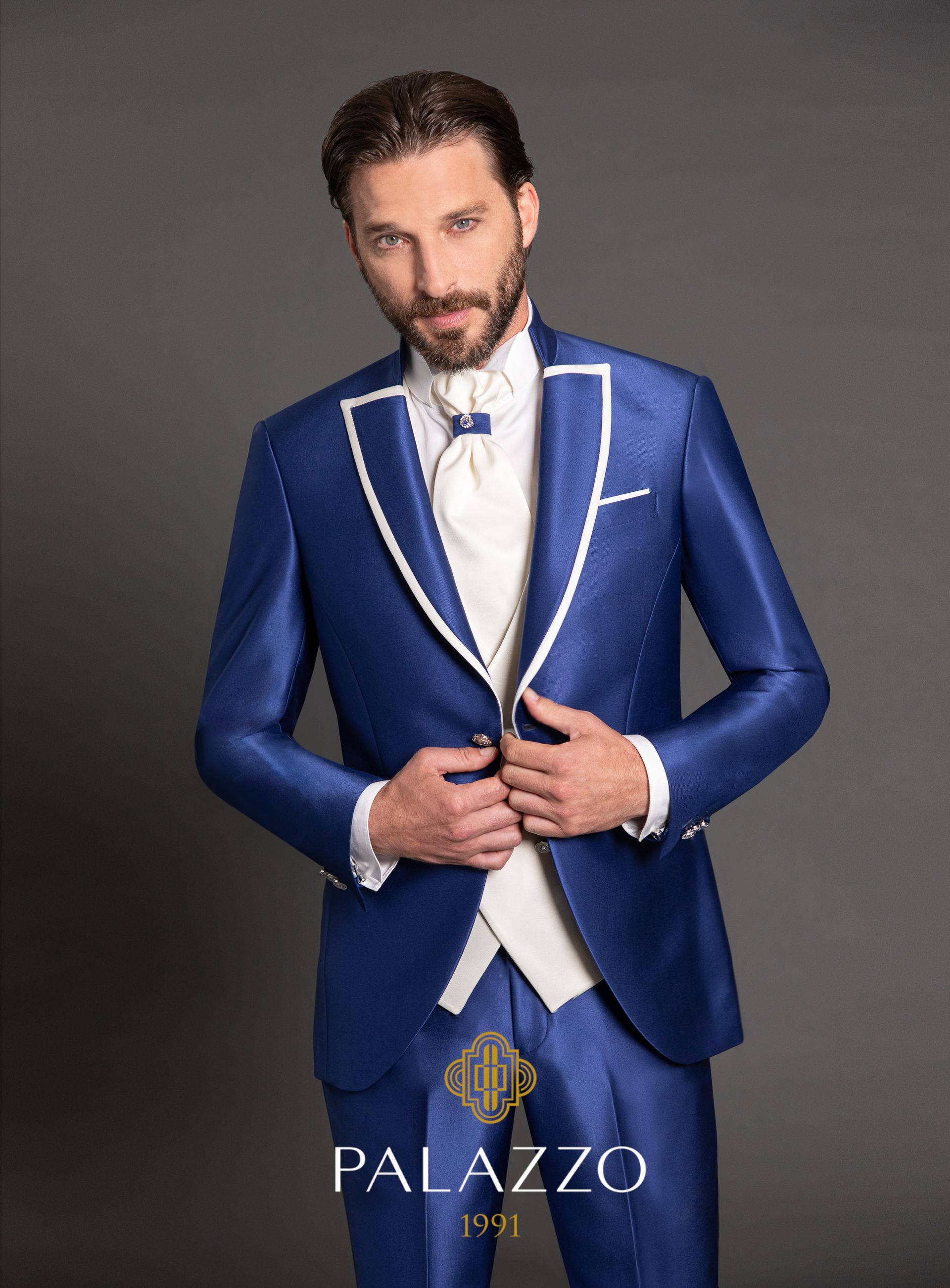 Uomo in abito blu con rifiniture bianche e gilet, che abbottona la giacca. Sfondo grigio.