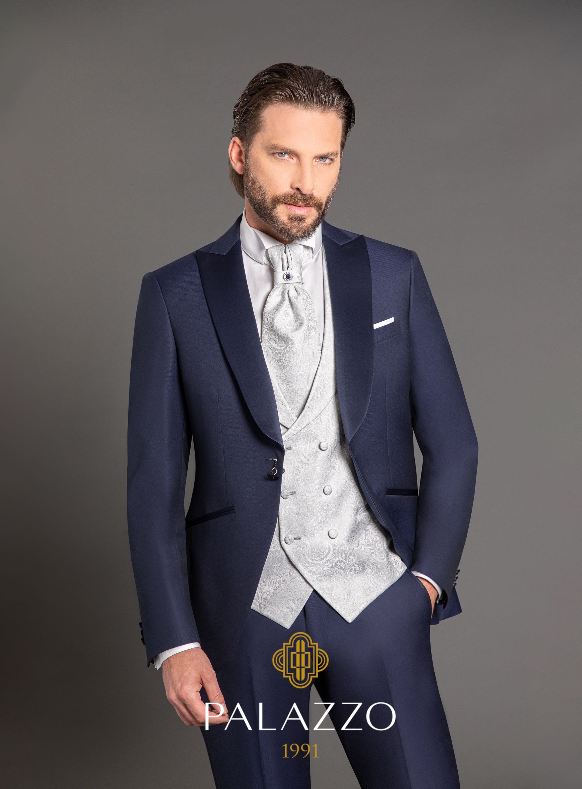 Uomo in smoking blu navy con gilet con fantasia argentata, in posa su uno sfondo grigio.