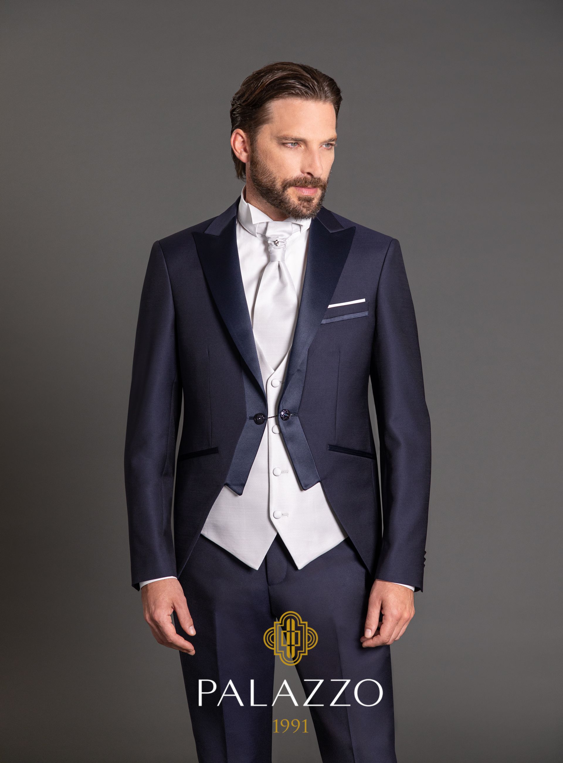 Uomo in abito formale blu navy con gilet leggero e cravatta, in piedi su uno sfondo grigio.