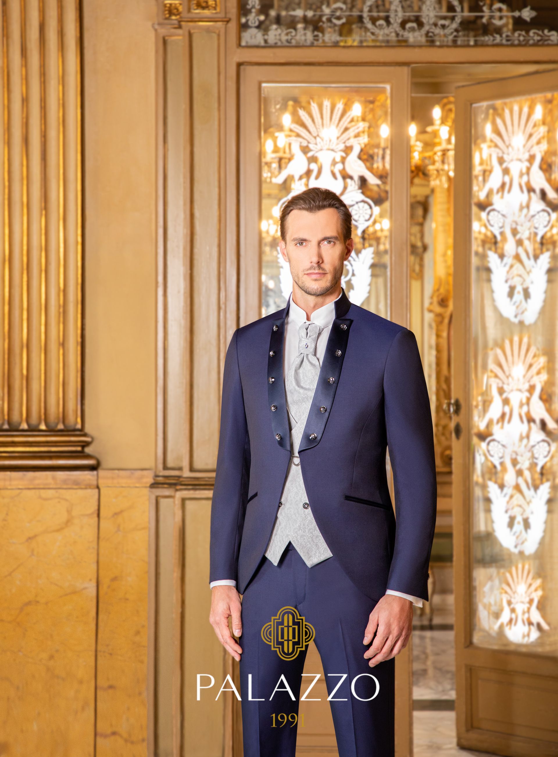 Uomo in abito blu, gilet e camicia bianca, in piedi sulla soglia decorata. È visibile il logo del Palazzo.