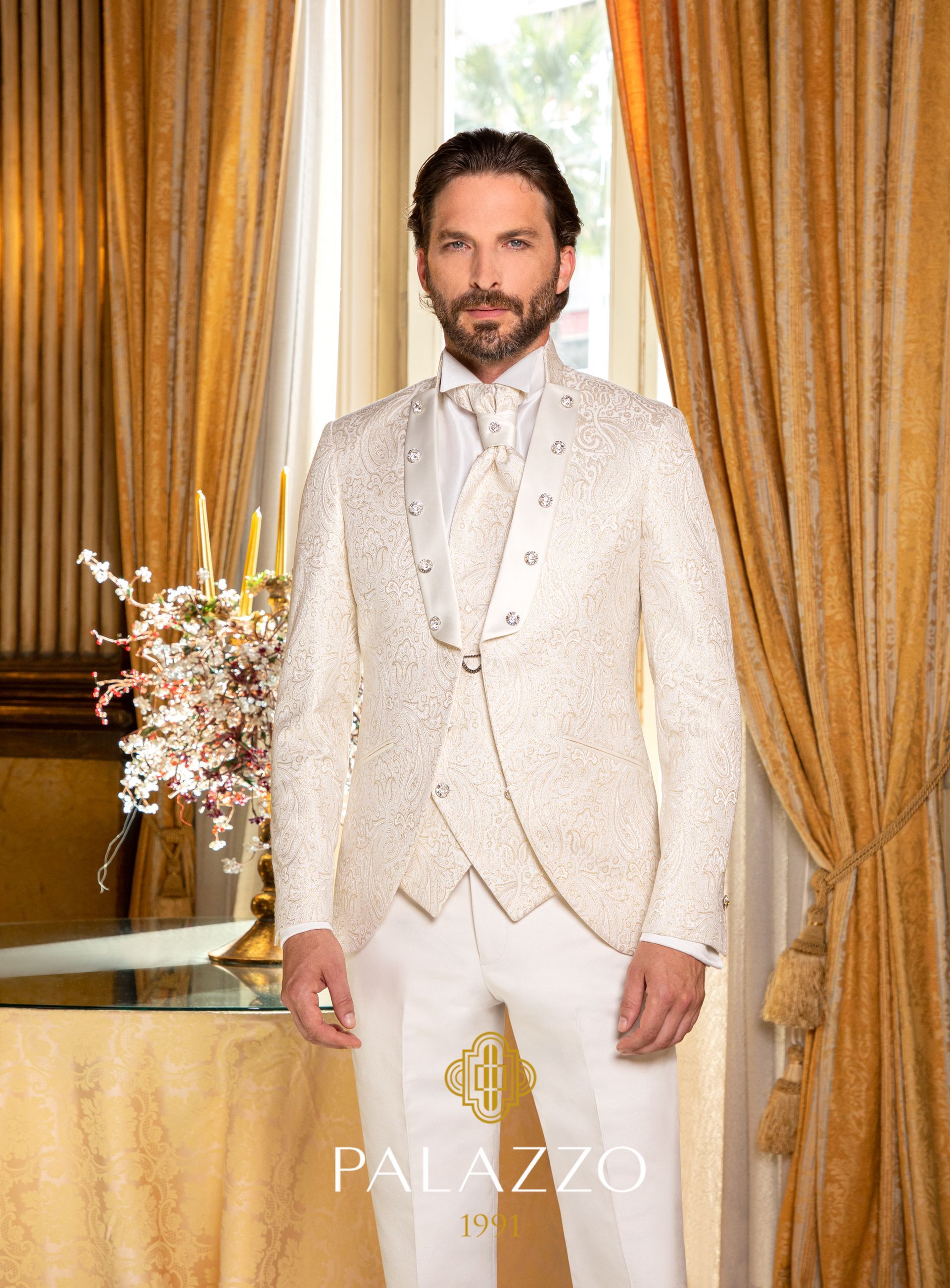 Un uomo in un abito color crema testurizzato è in piedi all'interno. Indossa un gilet e una cravatta con una giacca decorata.