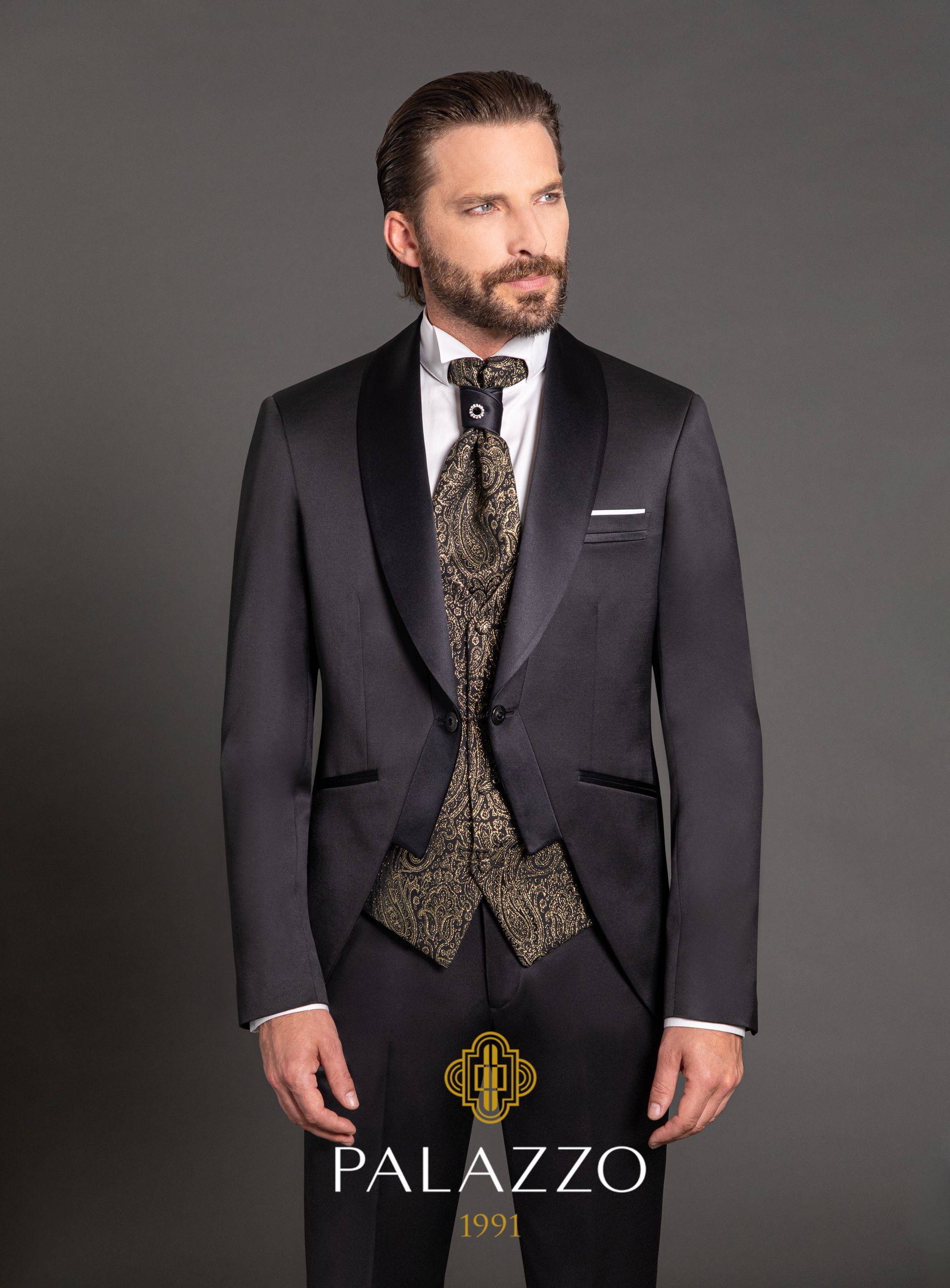 Uomo in abito nero formale, gilet con motivo dorato e cravatta, che guarda altrove, in uno studio.