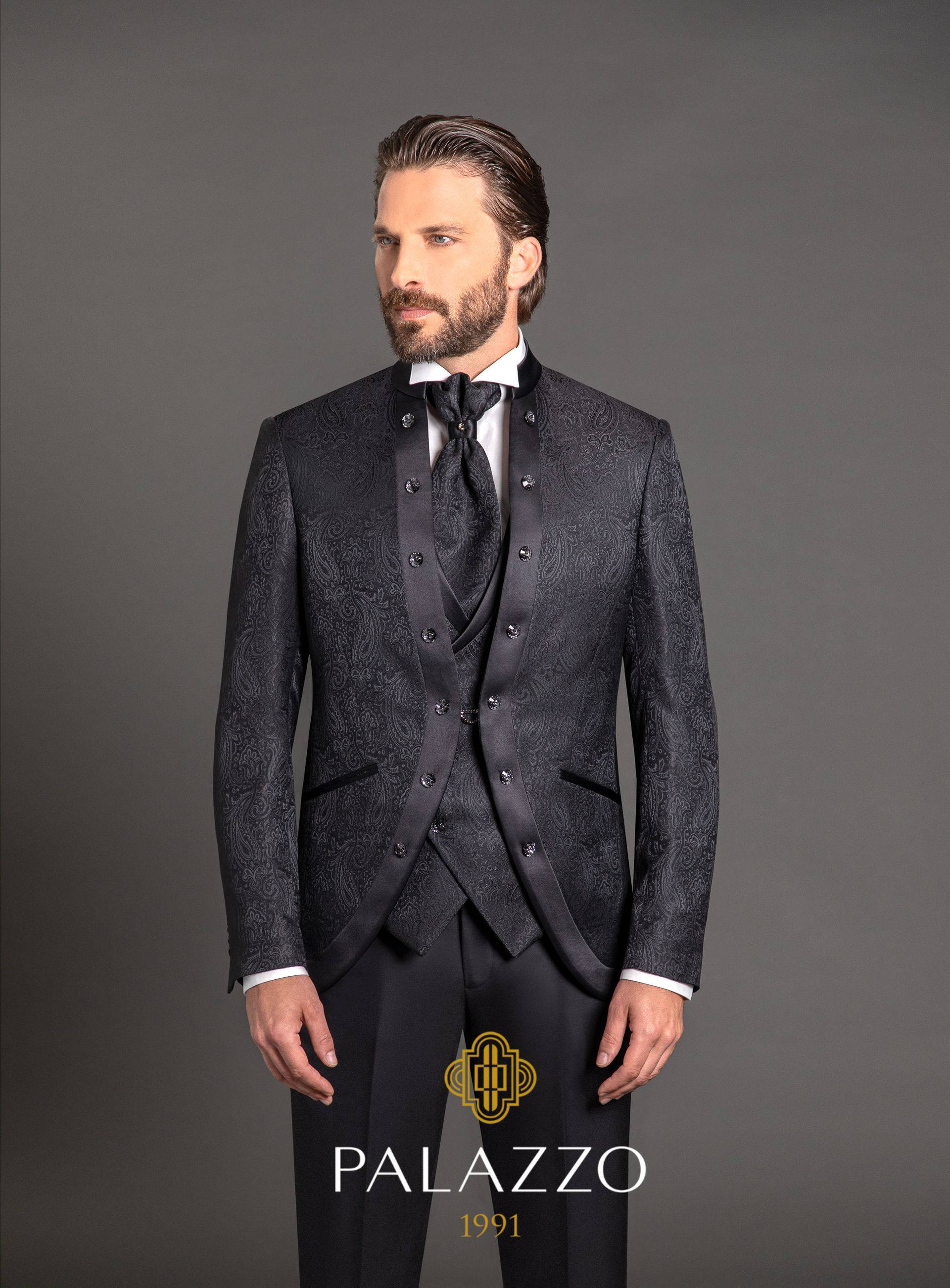 Uomo in un elegante abito nero con giacca fantasia e gilet coordinato, in piedi su uno sfondo grigio.