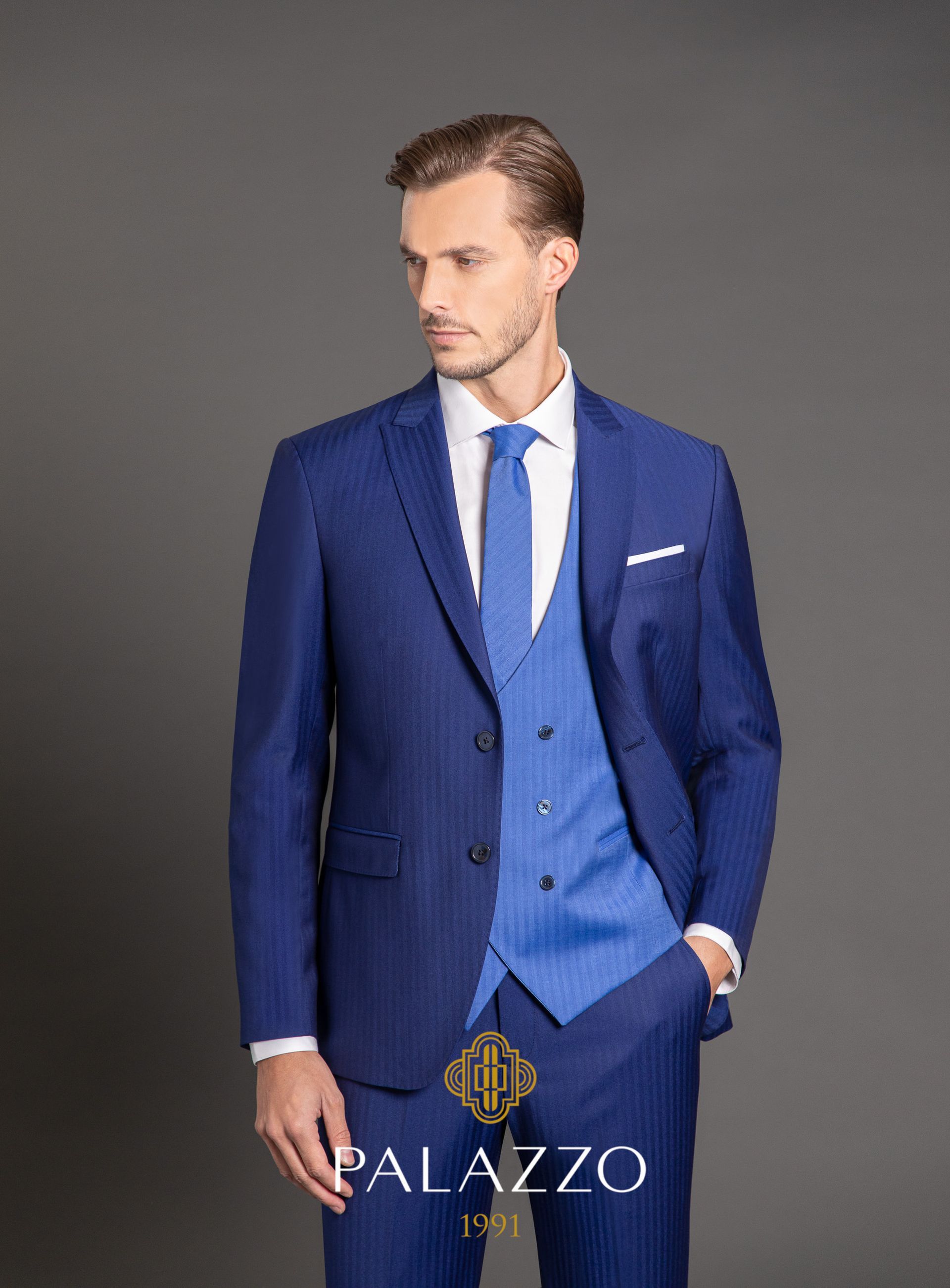 Uomo in abito blu, gilet e cravatta, che guarda di lato su uno sfondo grigio.