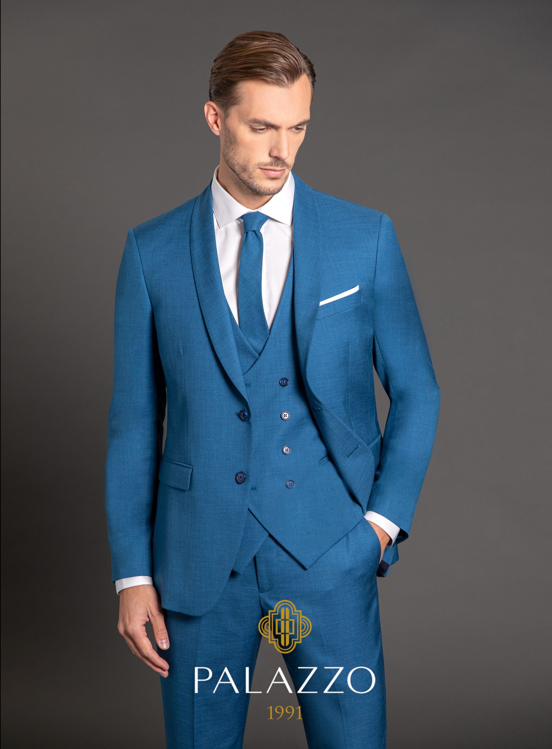 Uomo in abito blu con gilet, camicia bianca e cravatta, che guarda in basso con una mano in tasca, su uno sfondo grigio.