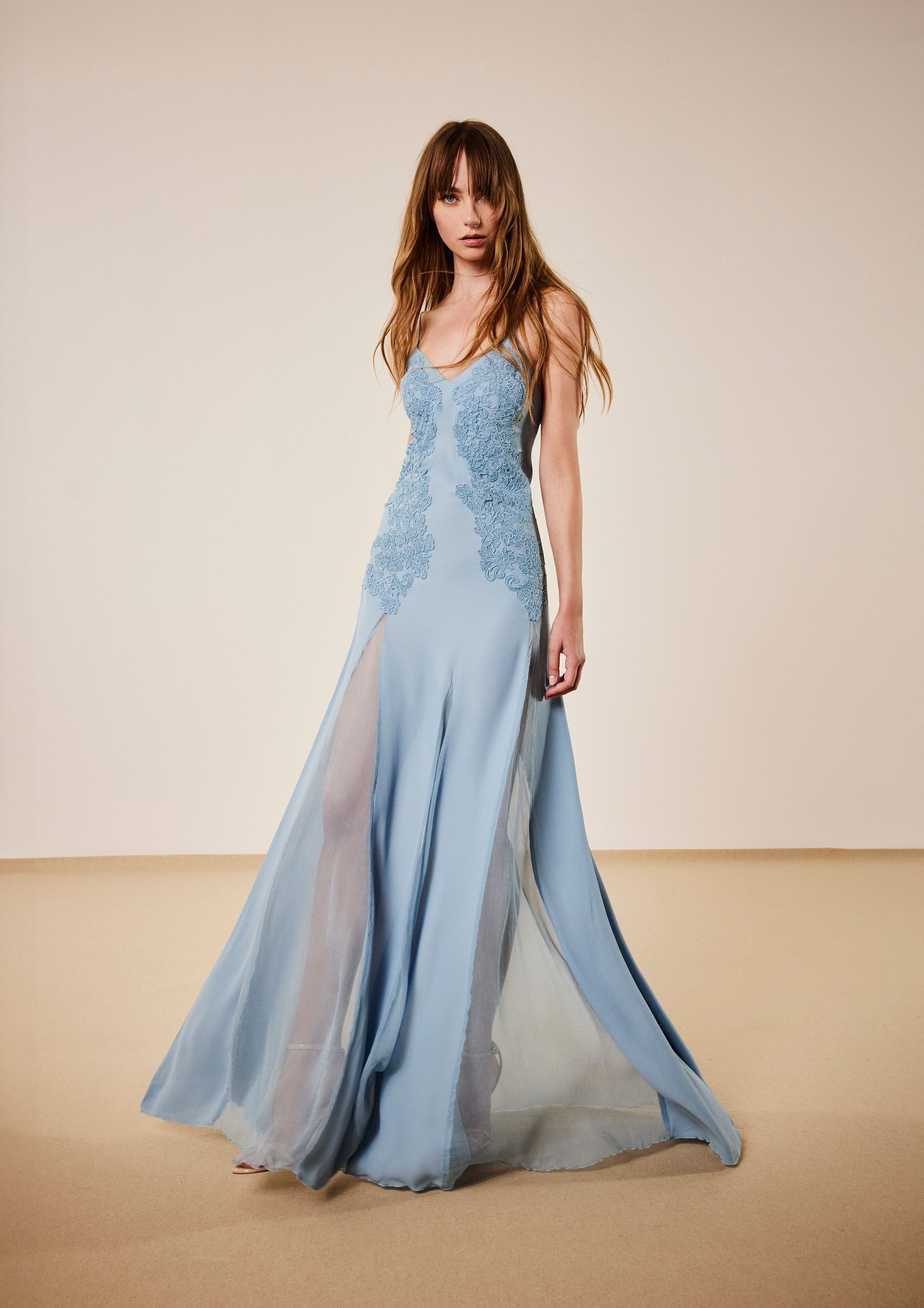 Donna in abito da sera blu con decorazioni floreali, in piedi in uno studio con pareti dai colori chiari.