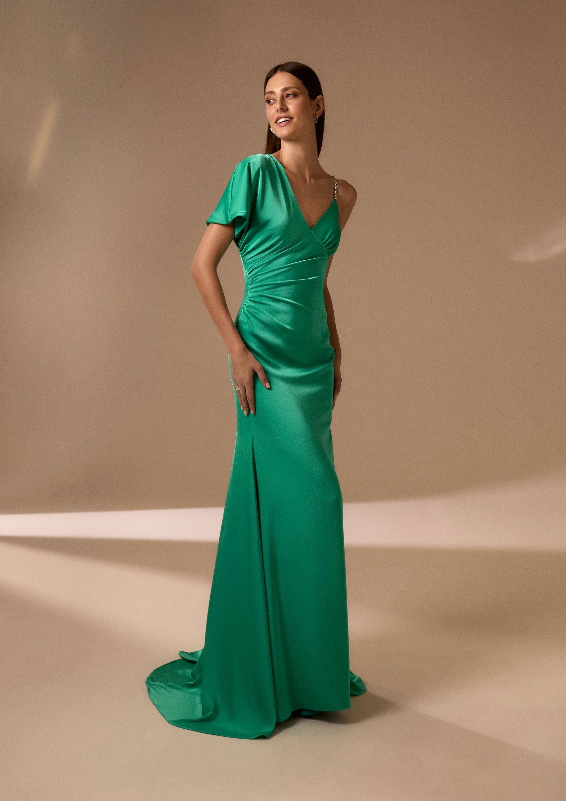 Donna in abito di raso verde con manica monospalla, in posa in un interno.