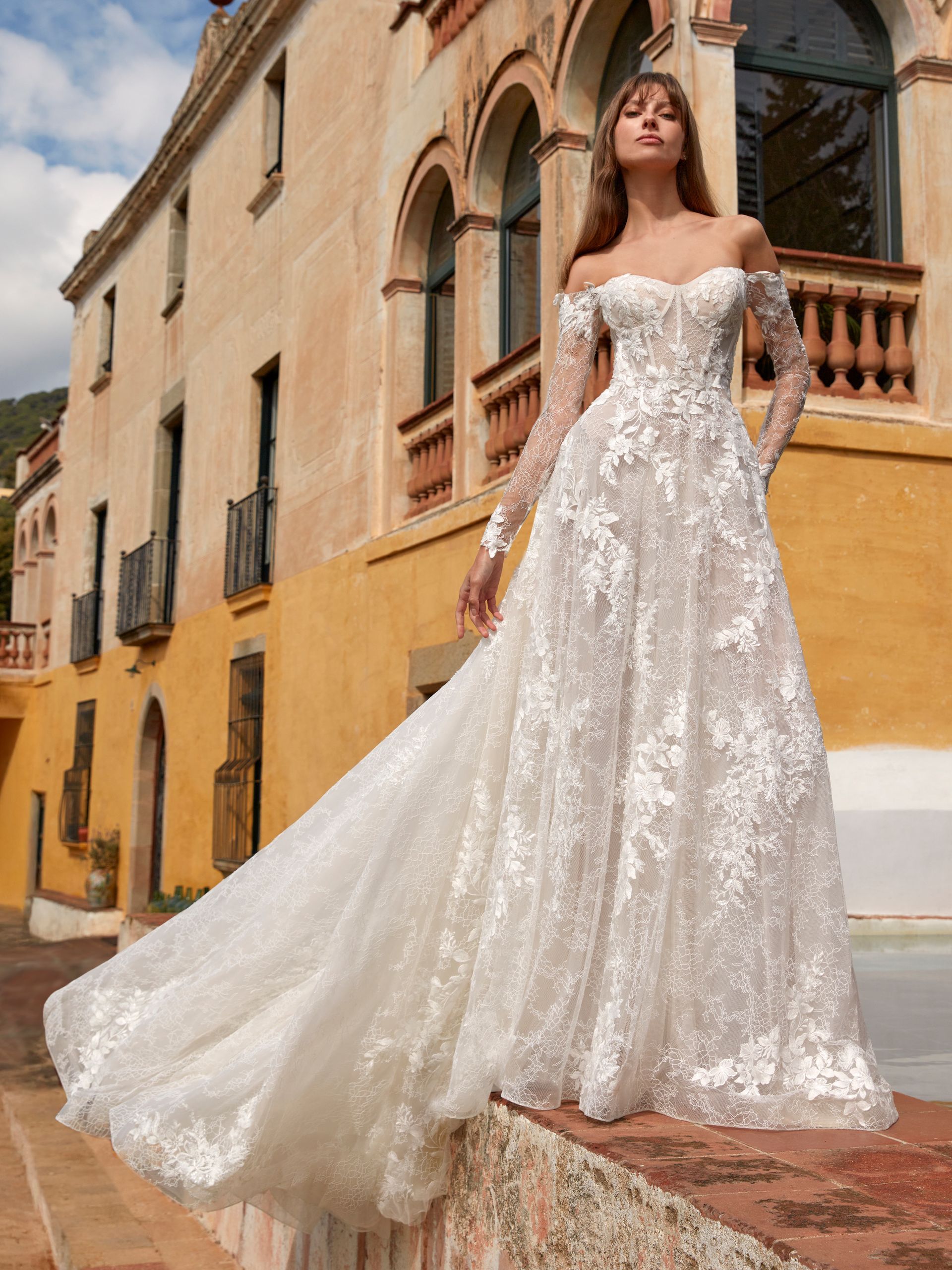 Donna in abito da sposa in pizzo bianco con spalle scoperte e lungo strascico, in piedi su una sporgenza 