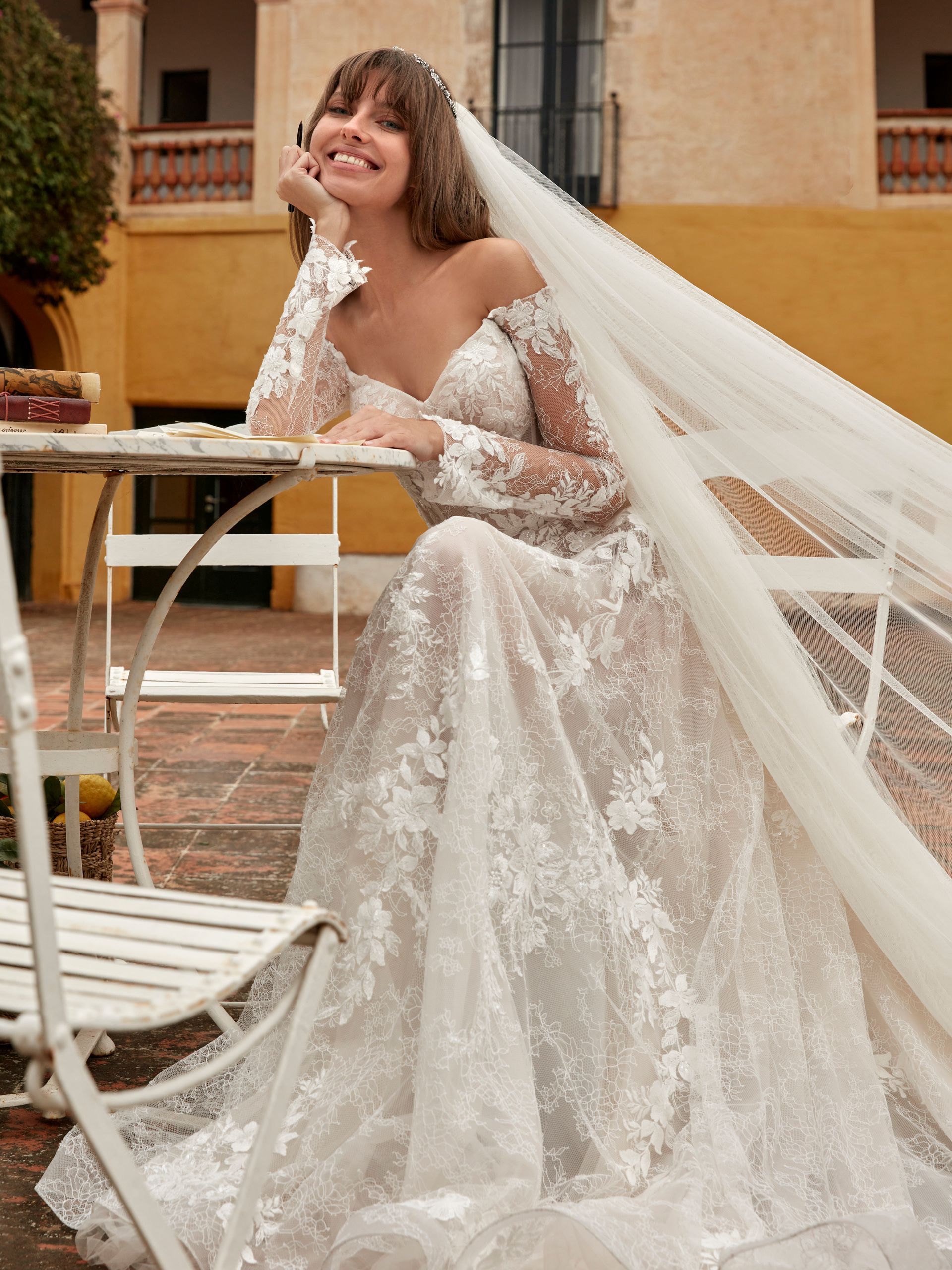 Sposa in abito da sposa in pizzo con spalle scoperte, sorridente, seduta a un tavolo all'aperto, con il velo svolazzante.