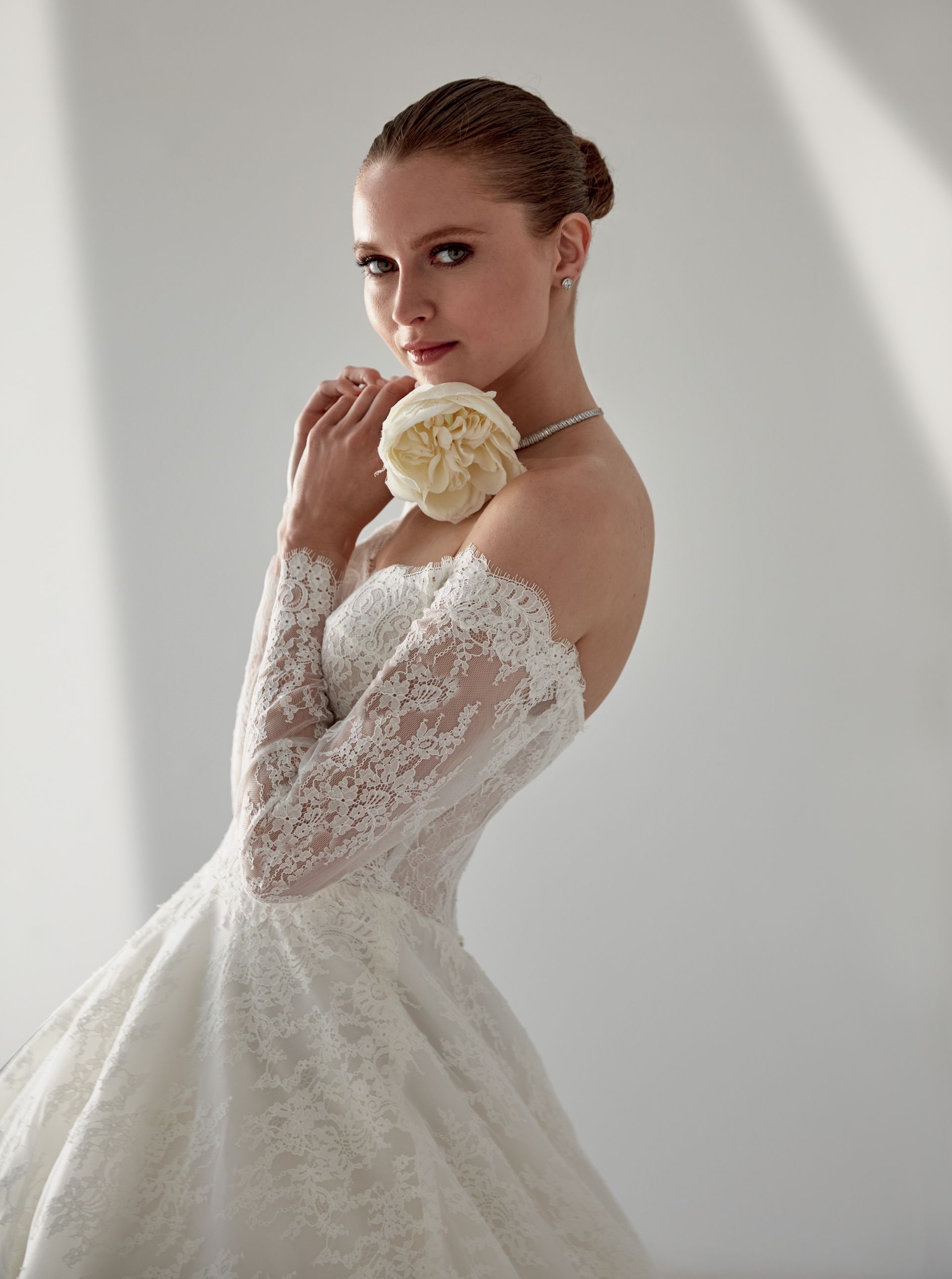 Donna con abito da sposa in pizzo bianco, che tiene in mano un fiore, sorridente, su uno sfondo bianco.