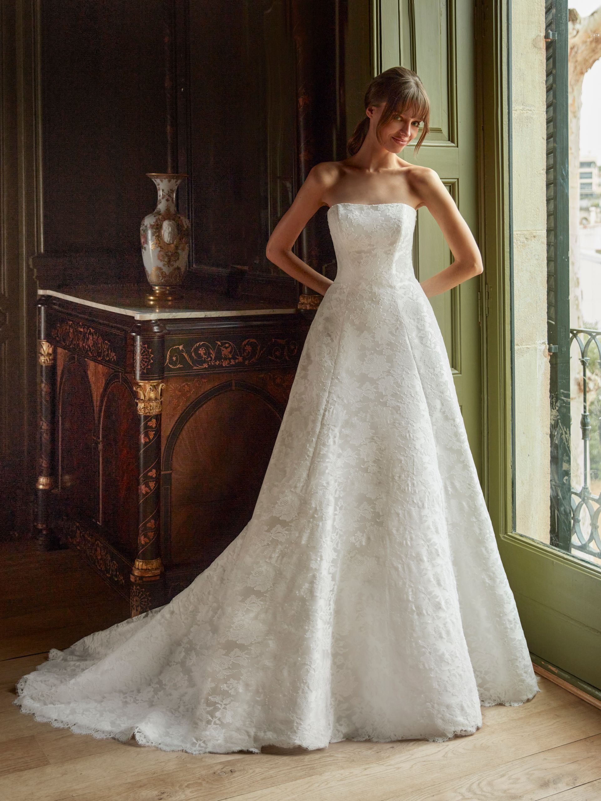 Una donna in abito da sposa bianco senza spalline è in piedi vicino a una finestra in una stanza.