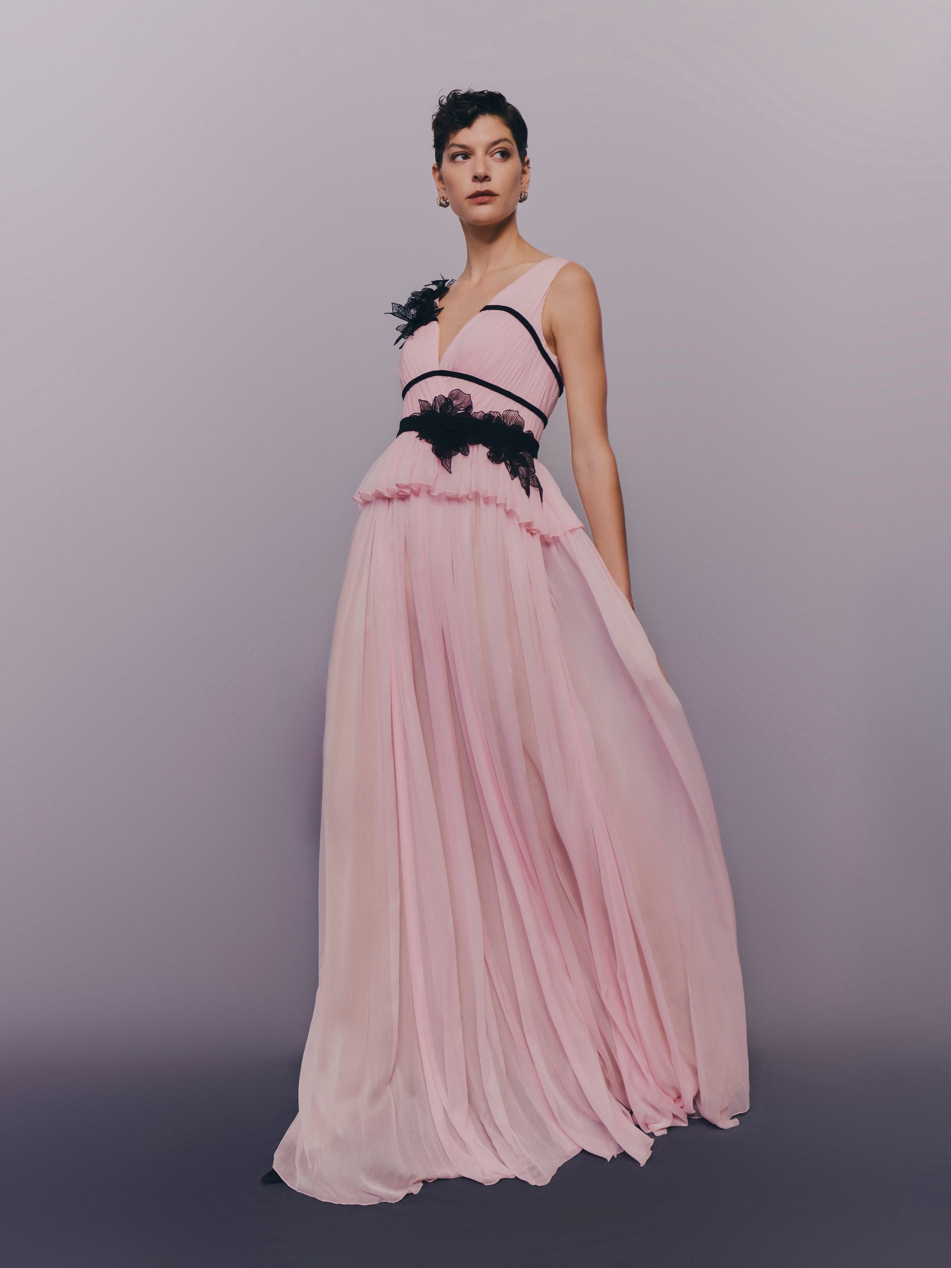 Donna in abito lungo rosa con dettagli in pizzo nero in posa su uno sfondo neutro.