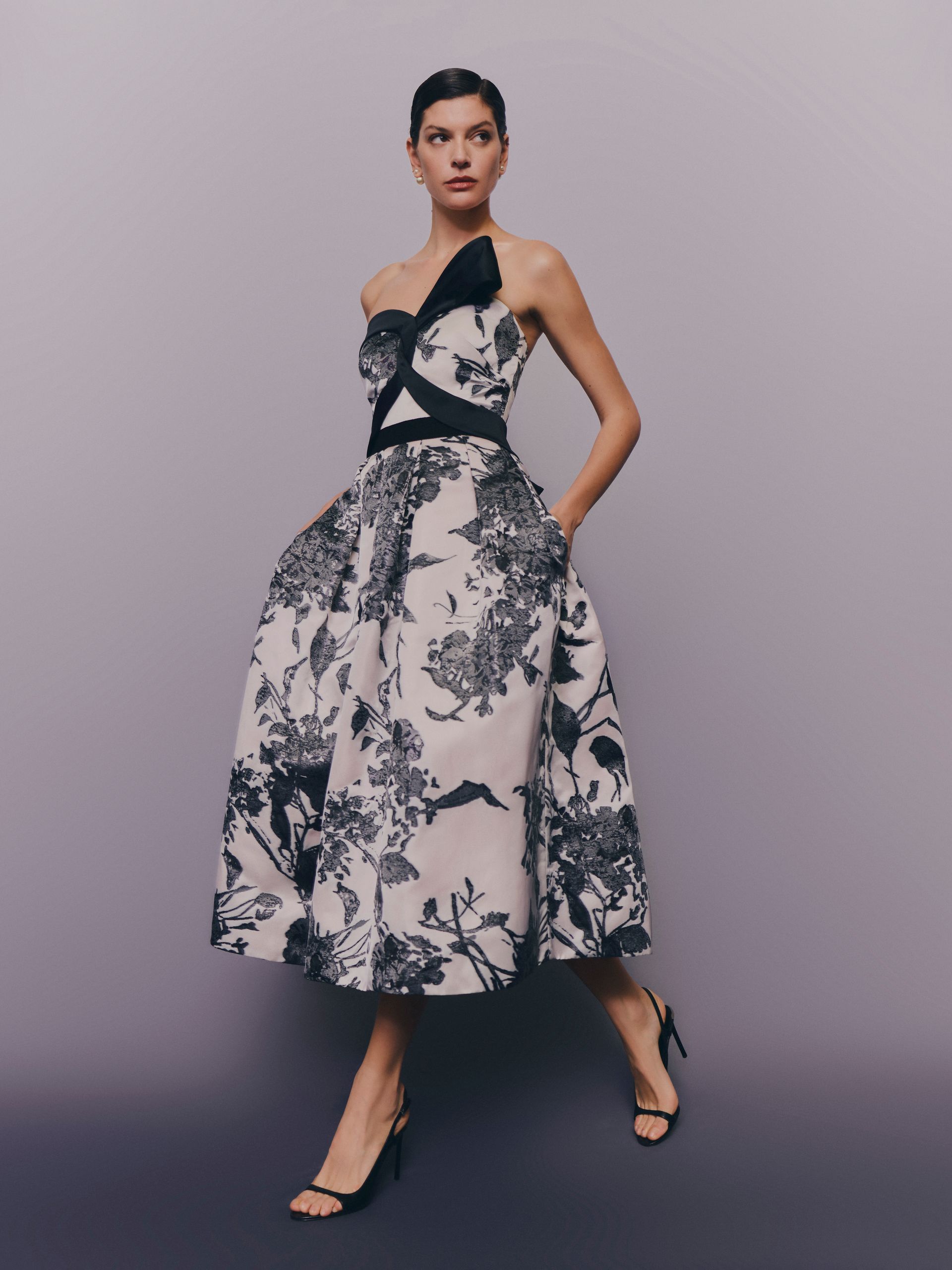 Donna in un abito monospalla floreale con dettagli neri e tacchi, in piedi in uno studio.