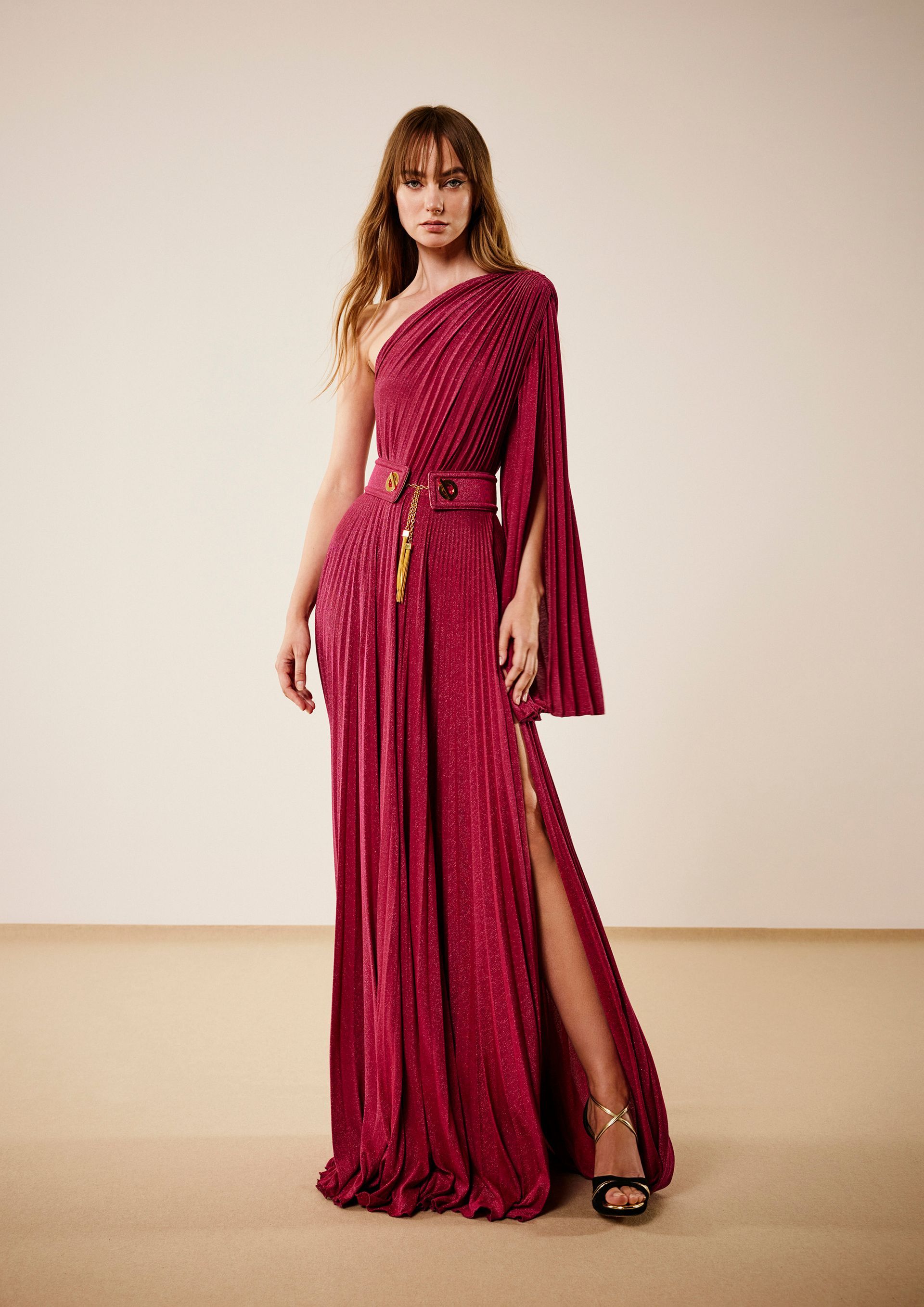 Donna in abito rosso monospalla con manica aperta e spacco profondo, in posa davanti a uno sfondo color crema.