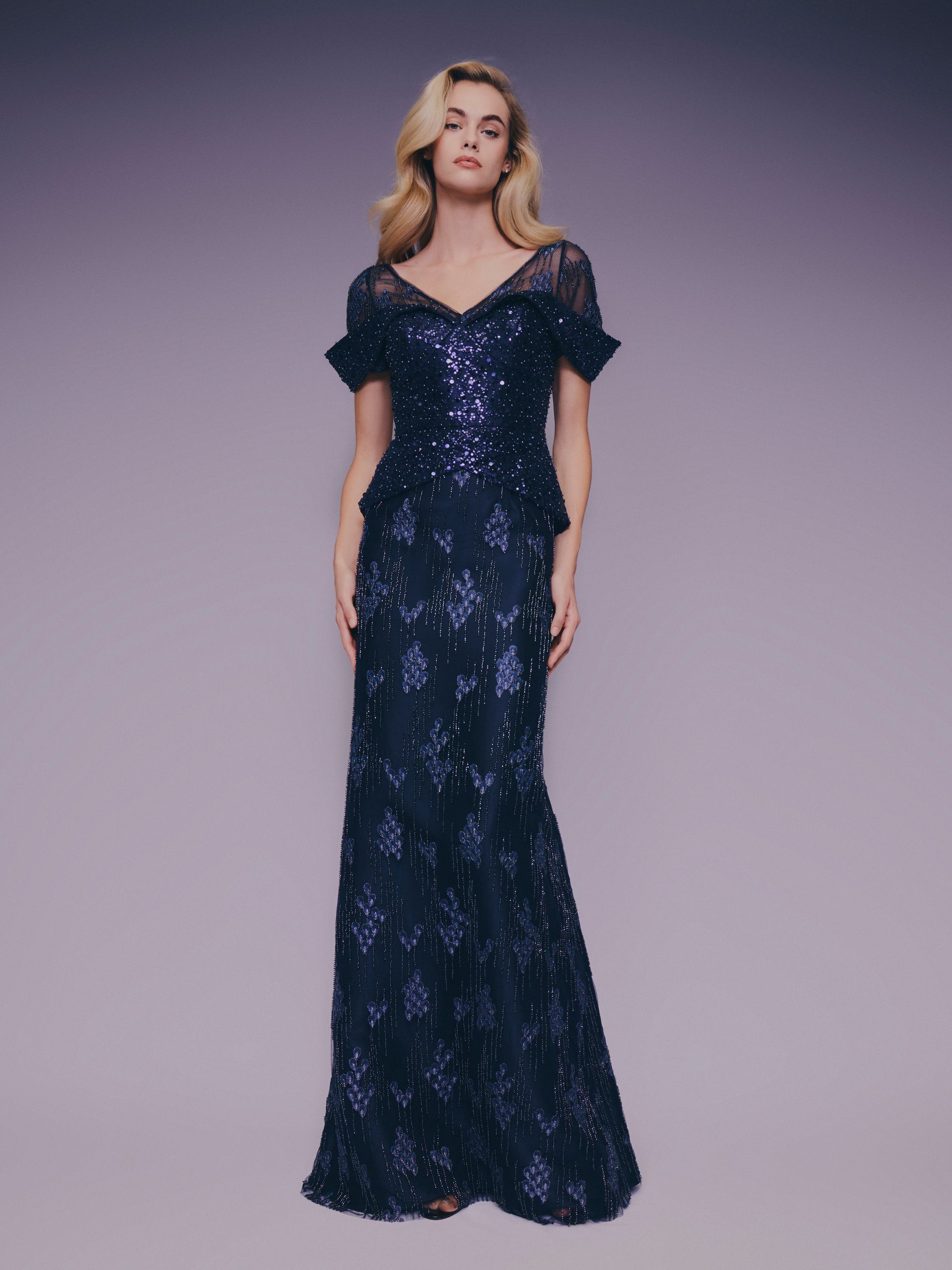 Donna in abito da sera blu scuro con paillettes, in piedi su uno sfondo viola.