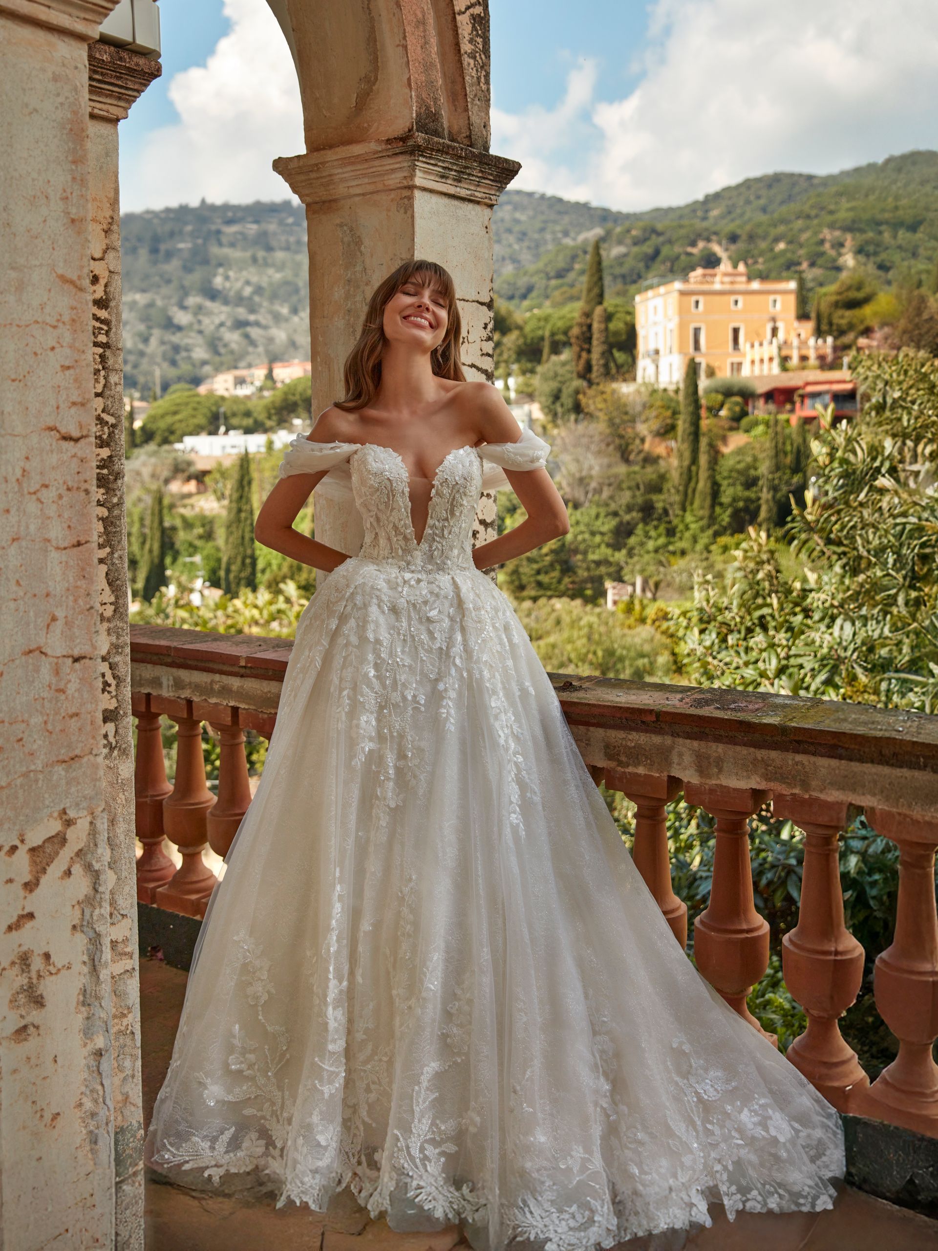 Donna in abito da sposa con spalle scoperte sul balcone con montagne e palazzi sullo sfondo.
