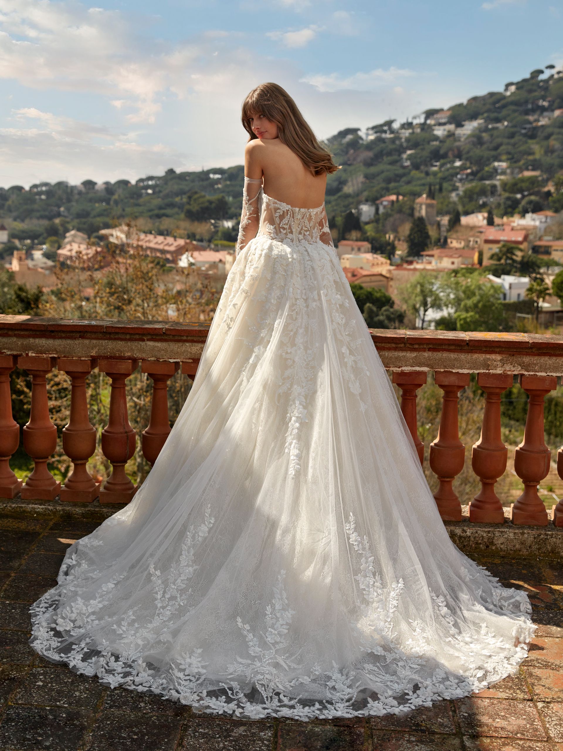 Donna in abito da sposa bianco con lungo strascico, in piedi su un balcone che si affaccia su un paesaggio pittoresco.