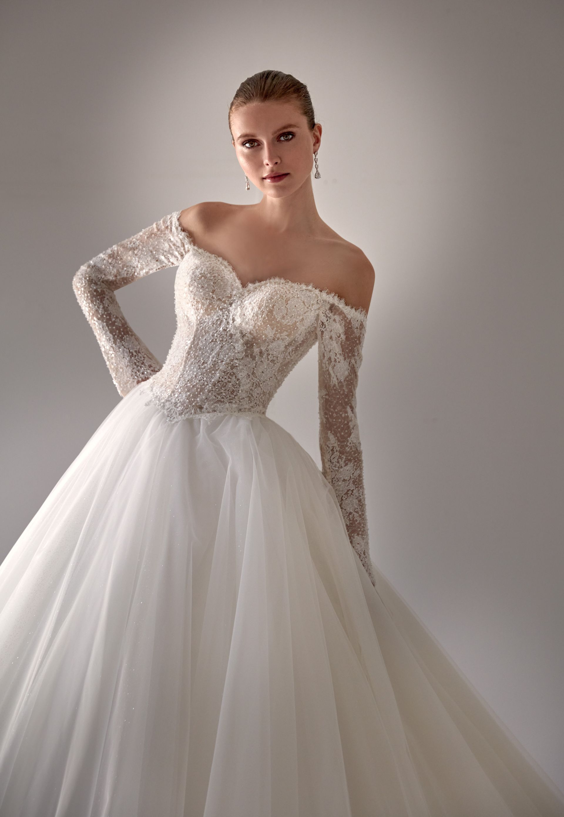 Donna in abito da sposa bianco con spalle scoperte, corpetto in pizzo, gonna ampia, maniche lunghe, in posa in un interno.