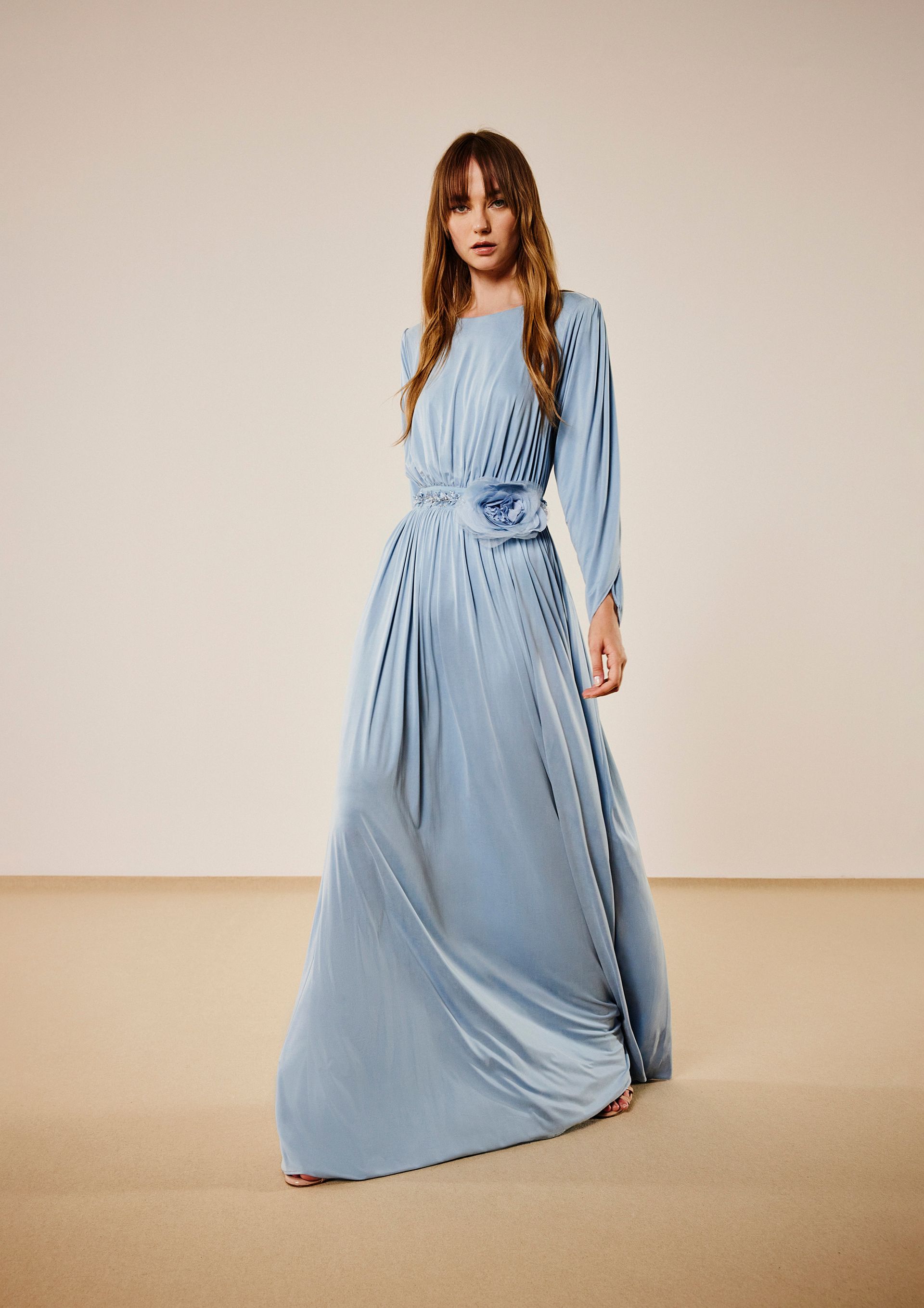 Donna con un lungo abito azzurro plissettato a maniche lunghe. Ha i capelli castano chiaro ed è in piedi in casa.