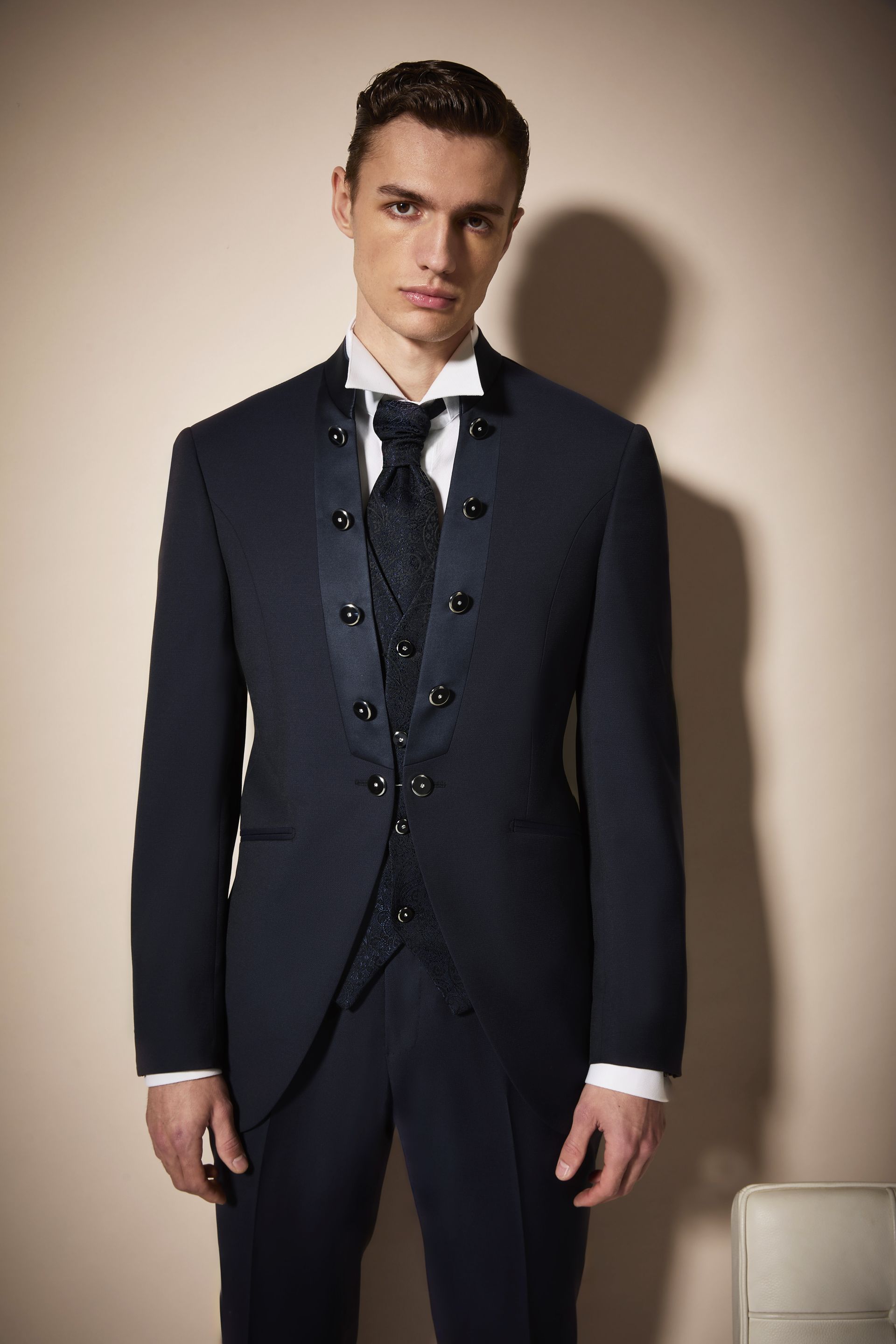Uomo in abito formale blu scuro con gilet, in piedi davanti a uno sfondo neutro.