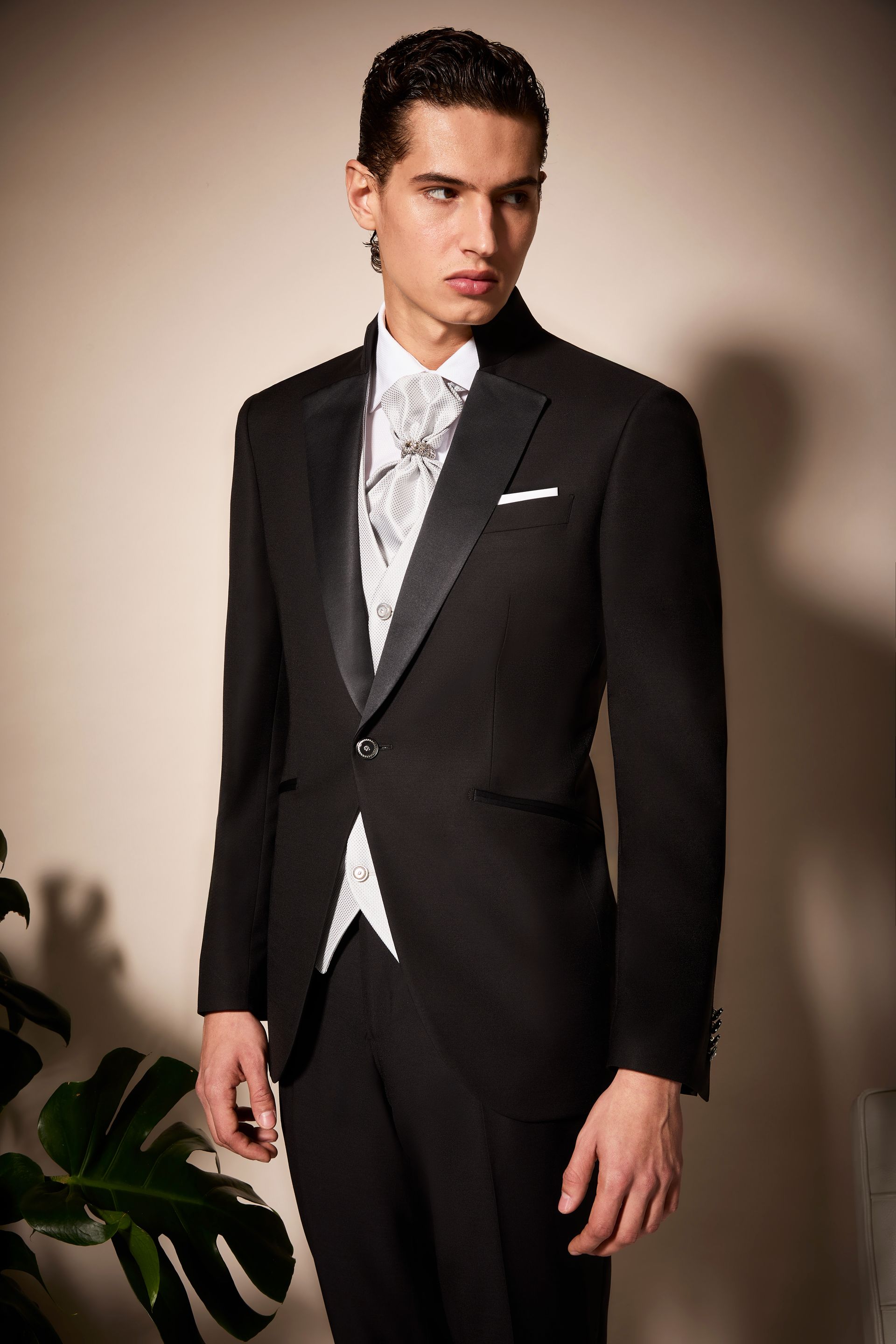Uomo in abito formale nero, gilet bianco fantasia, in posa in un interno.